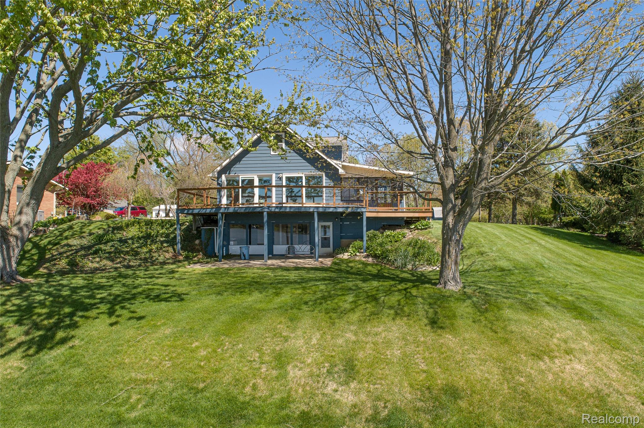 2855 Farmers Creek Road Metamora, MI 48455 - Photo 46 of 51 Blue siding exterior featuring a full-width upper deck with wooden railings, a lower-level patio with multiple glass doors, and a sloped, manicured lawn with mature trees