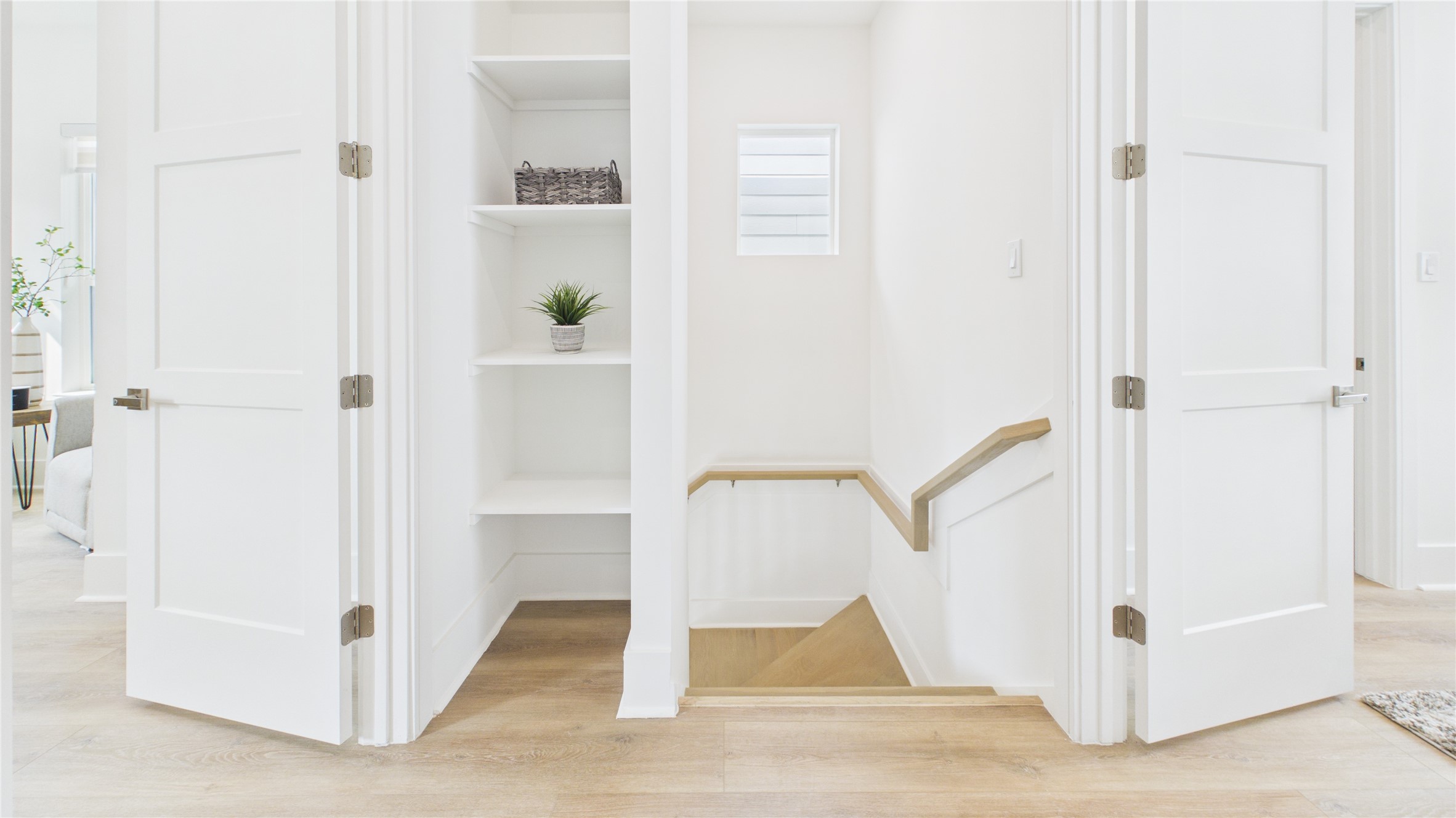 602 Skyland Terrace Way Houston, TX 77009 - Photo 20 of 40 This photo shows a bright, clean hallway featuring light wood floors, white walls, and doors. There are open shelves with decorative items, and a staircase with a wooden railing leading down. The space is modern and inviting, with natural light from a small window.