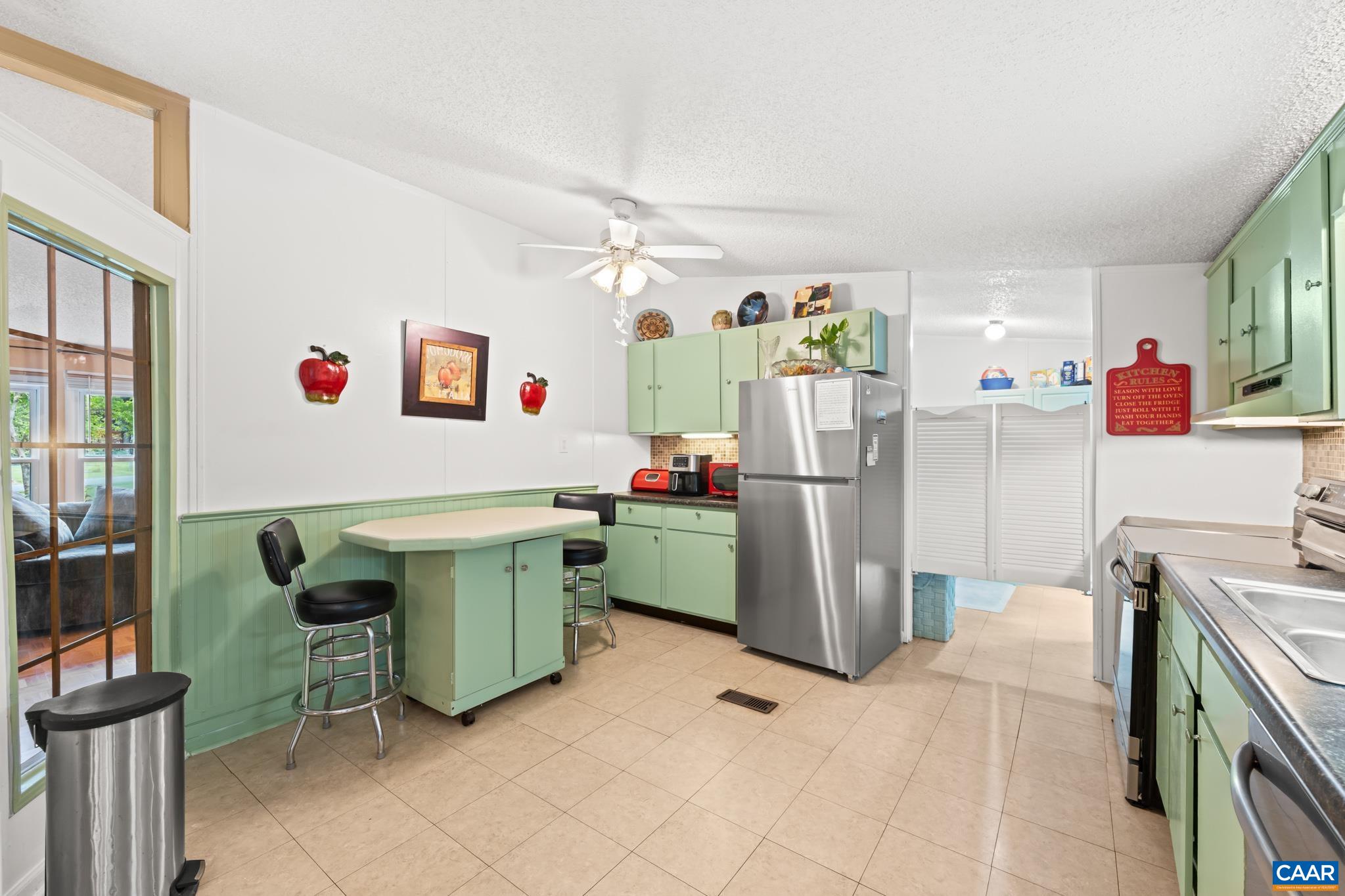 Spacious kitchen featuring retro sage green cabinetry, a central island with breakfast bar seating, stainless steel refrigerator, and ample prep space. Complete with a ceiling fan and charming accent details, it's ideal for casual dining!