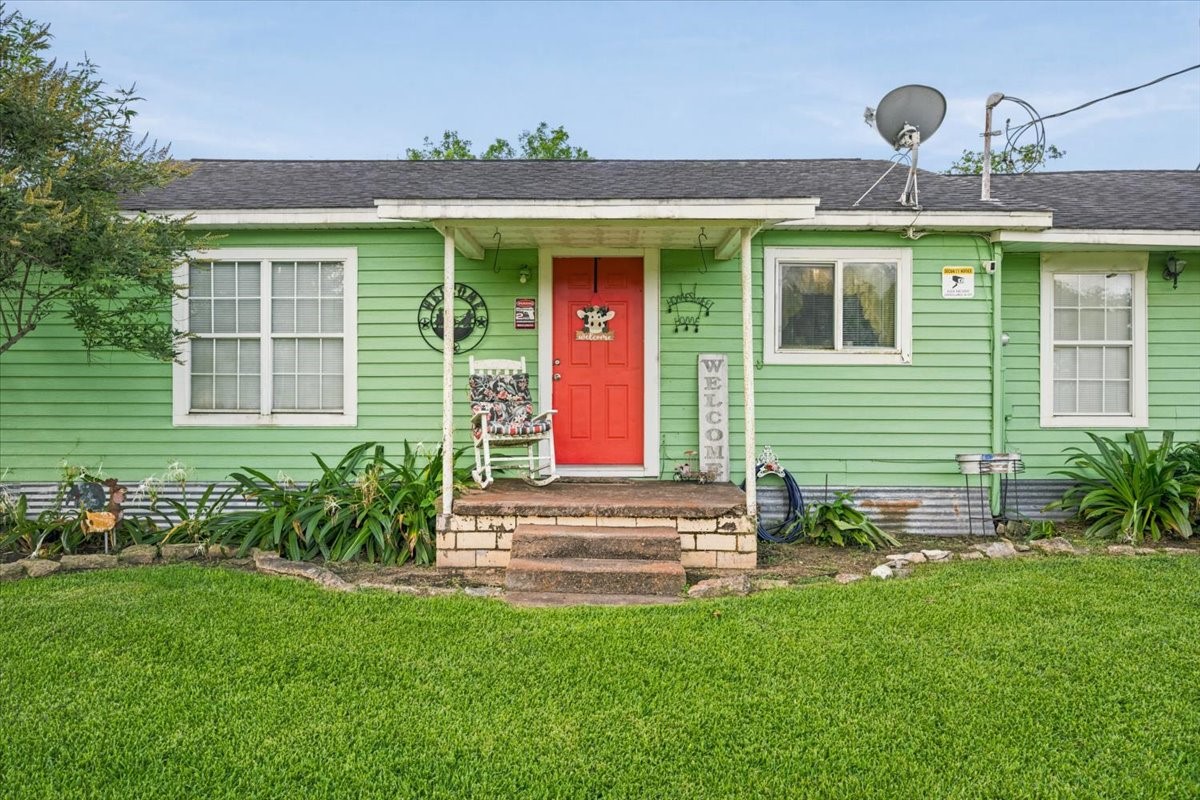765 Norcross Road Wallis, TX 77485 - Photo 2 of 42 Charming single-story home with a vibrant green exterior and welcoming red front door. Features a cozy porch with a swing, surrounded by well-maintained landscaping and lush greenery.