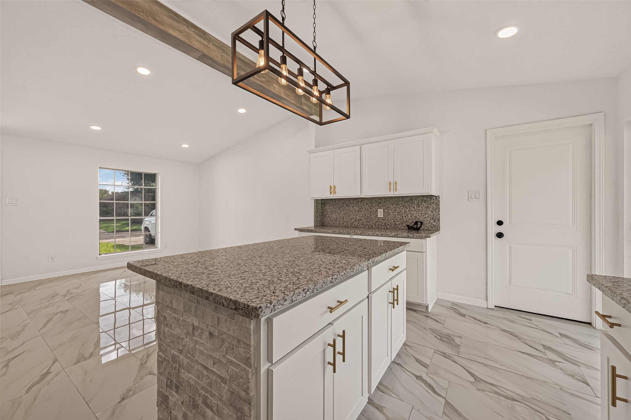 16619 Tibet Road Friendswood, TX 77546 - Photo 9 of 29 This photo showcases a modern kitchen with sleek granite countertops, white cabinetry, and stylish gold hardware. The open layout features a central island with a brick accent, under a contemporary light fixture. The space is bright with natural light from a large window, complemented by polished tile flooring.