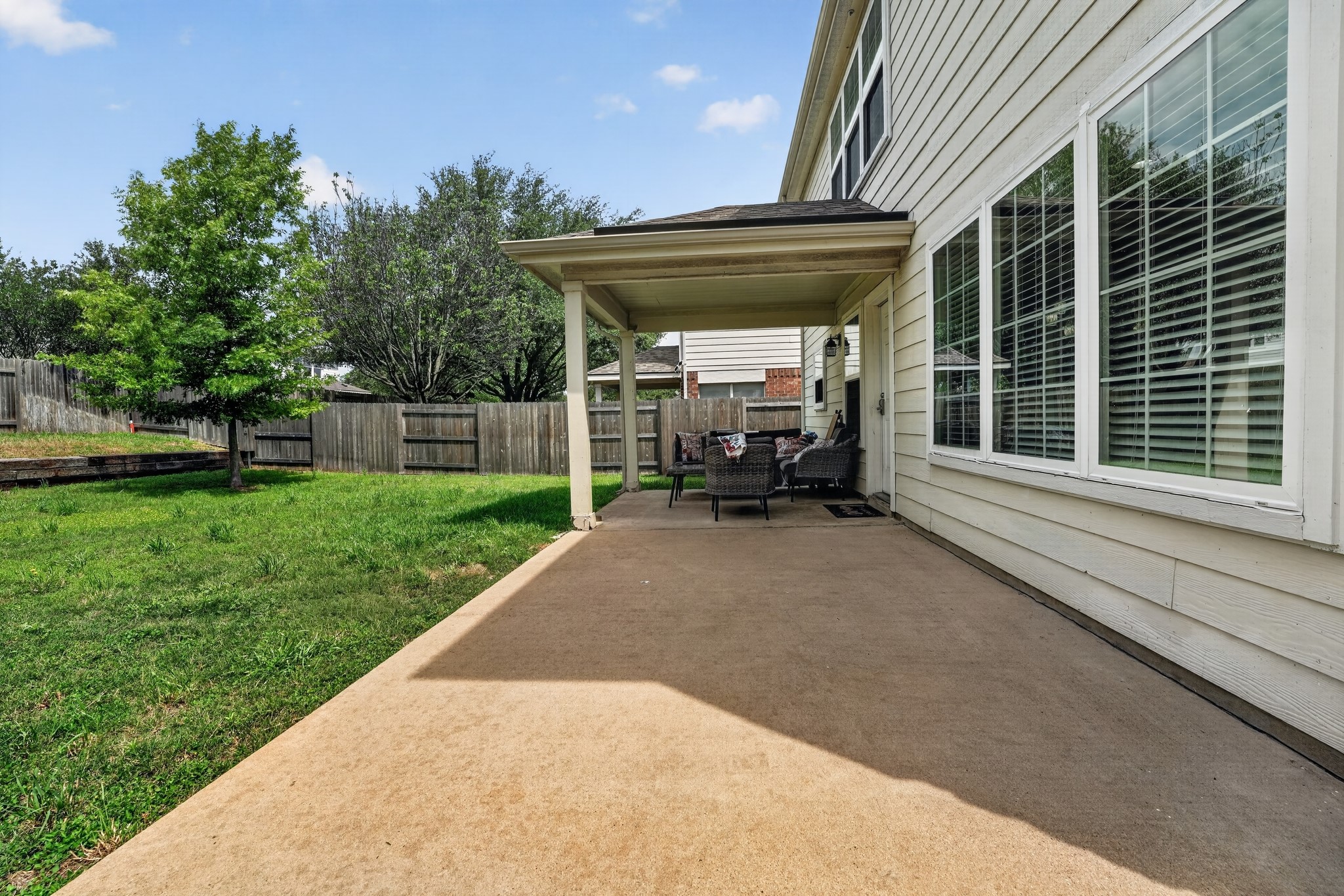 Expansive concrete patio offering a covered area with support columns, featuring large windows with blinds, and a durable siding exterior
