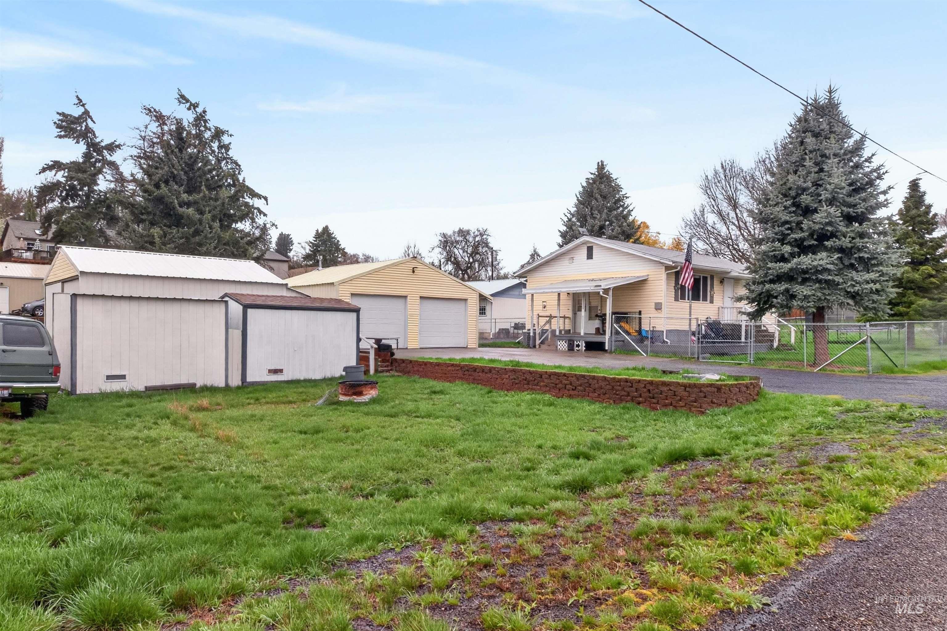 247 North Beech Street Genesee, ID 83832 - Photo 3 of 27 Expansive property featuring a single-story residence with a covered front porch, a two-car garage, multiple storage sheds, and mature evergreen trees