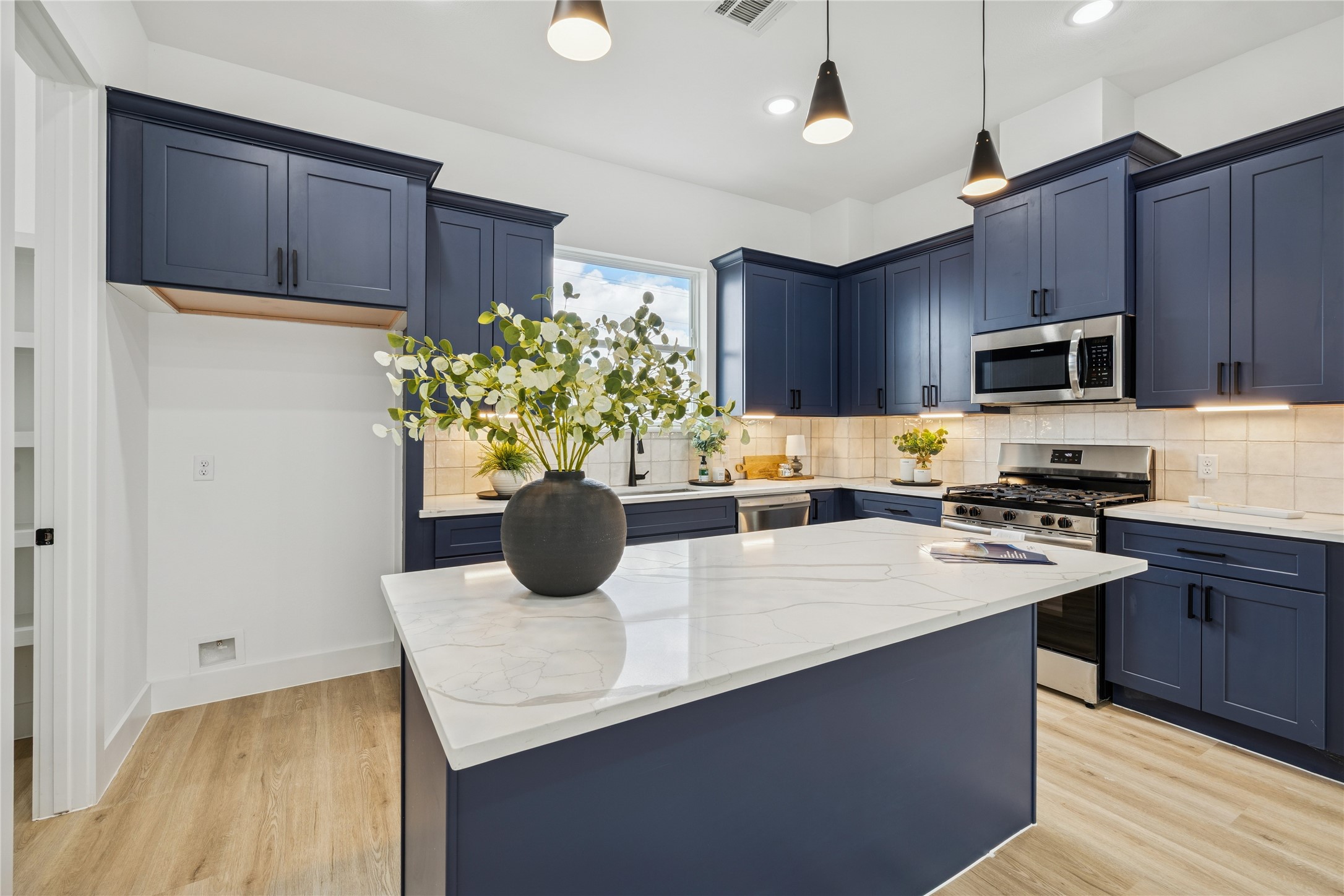 111 Milby Street Houston, TX 77003 - Photo 14 of 39 This kitchen features sleek navy cabinets, a modern marble island with a vase centerpiece, stainless steel appliances, and warm wood flooring, offering a stylish and functional space.