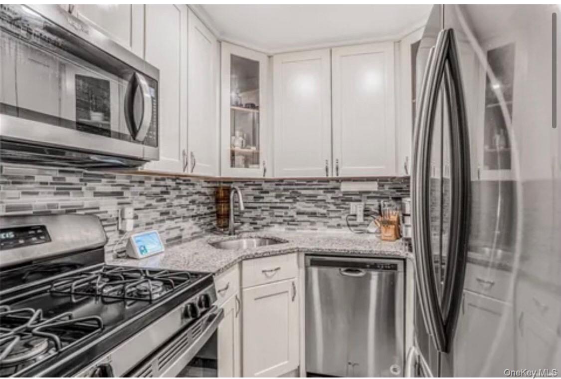 46-12 161 Street, Unit 2C Queens, NY 11358 - Photo 5 of 9 Kitchen with appliances with stainless steel finishes, glass insert cabinets, light stone countertops, white cabinets, and decorative backsplash