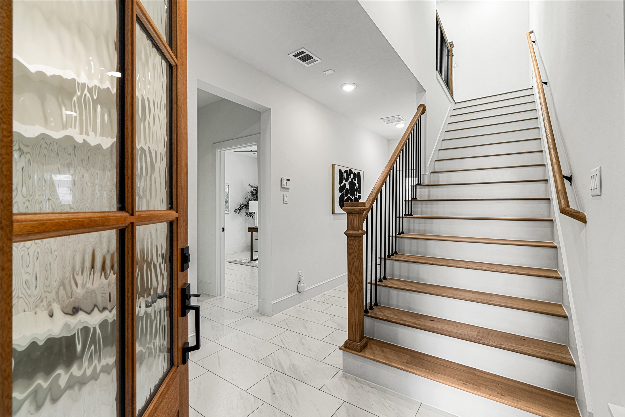 1614 Ojeman Road, Unit E Houston, TX 77055 - Photo 5 of 50 A bright entryway with a wooden front door, leading to a staircase with wooden steps and a metal railing. The space features white walls, tiled floors, and modern decor, creating a welcoming and contemporary atmosphere.