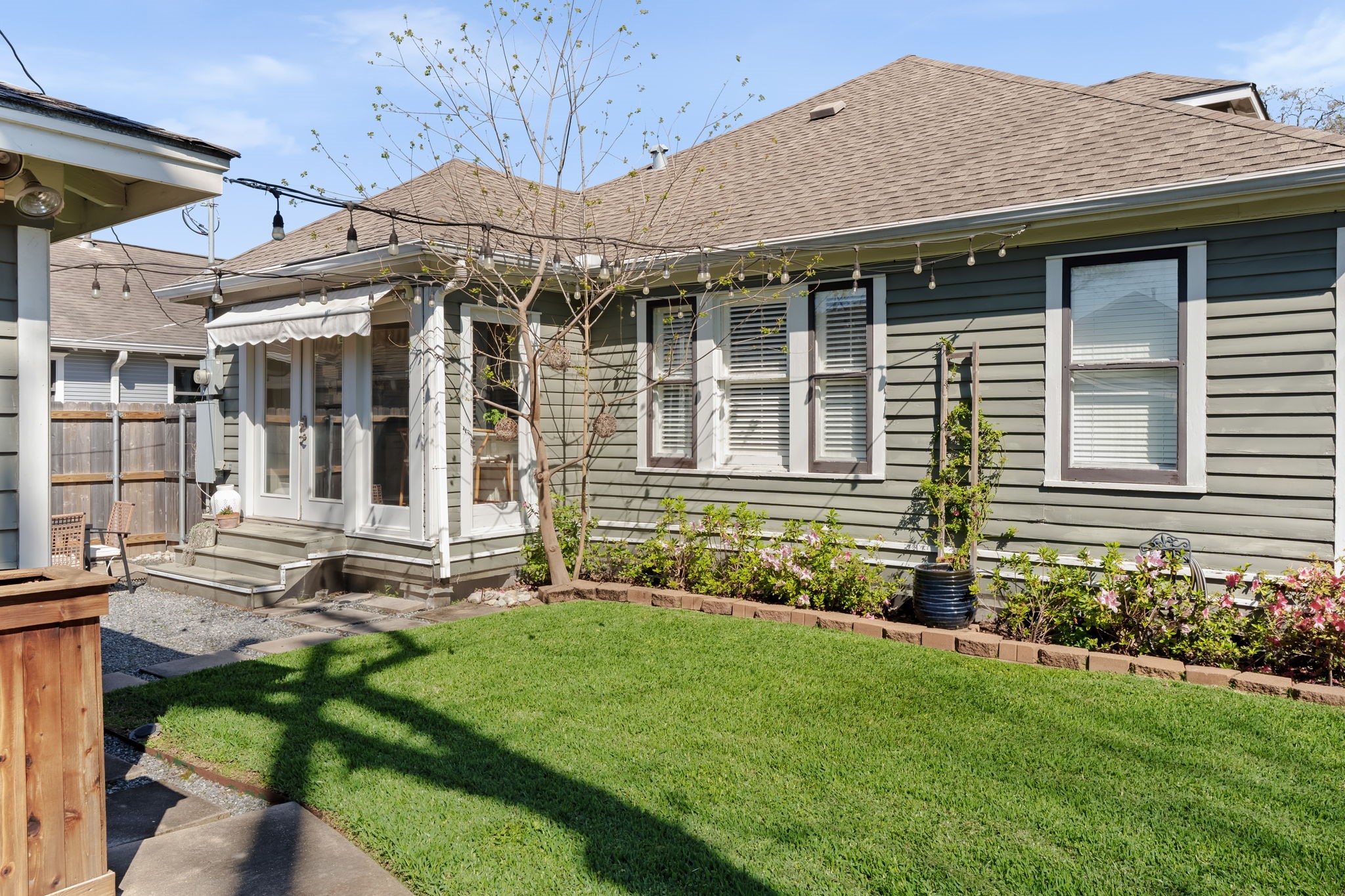 1002 West Cottage Street Houston, TX 77009 - Photo 26 of 37 This view back toward the house shows the kitchen bump-out with its awning-covered French doors, mature landscaping along the foundation, and string lights overhead. The yard is fully fenced with a wood privacy fence and gets good sun exposure throughout the day. It's a comfortable outdoor space for pets, kids, or just unwinding after work.