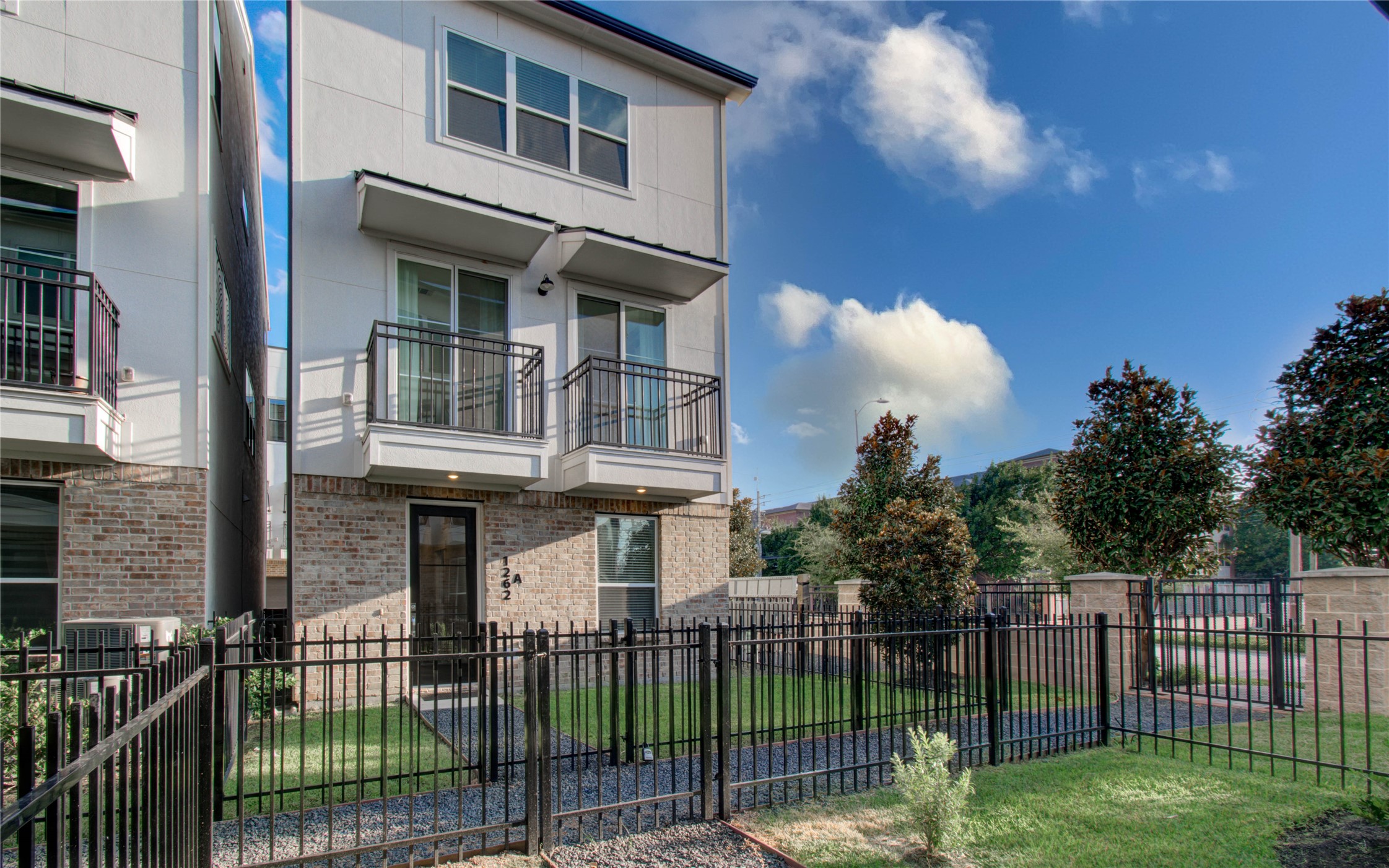 1262 North Post Oak Road, Unit A Houston, TX 77055 - Photo 3 of 50 This angled perspective highlights the front facade's dual second-floor balconies set above a cozy brick entry area. The fully fenced private front yard offers a perfect mix of lush turf, gravel paths, and young trees under a clear sky.