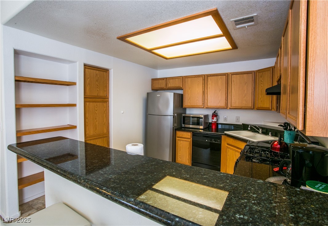 4655 Gold Dust Avenue, Unit 206 Las Vegas, NV 89120 - Photo 10 of 34 Kitchen with brown cabinetry, stainless steel appliances, a textured ceiling, dark stone counters, and a peninsula