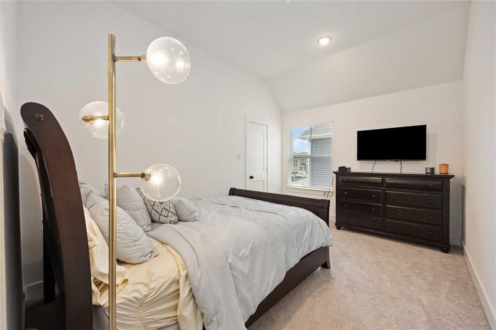 10014 Nectar Path Iowa Colony, TX 77583 - Photo 15 of 26 This cozy bedroom features a vaulted ceiling, soft carpeting, and a large window for natural light. It includes a modern floor lamp, a comfortable bed with plush bedding, and a dark wood dresser topped with a mounted TV, creating a relaxing ambiance.
