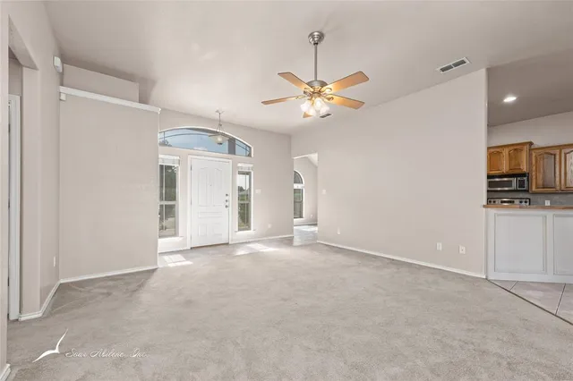 an empty room with chandelier fan and kitchen view