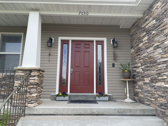 7030 West 2100 North Mendon, UT 84325 - Photo 5 of 67 Front entry featuring a red paneled door with decorative glass sidelights, stacked stone accents, gray horizontal siding, and integrated exterior lighting fixtures