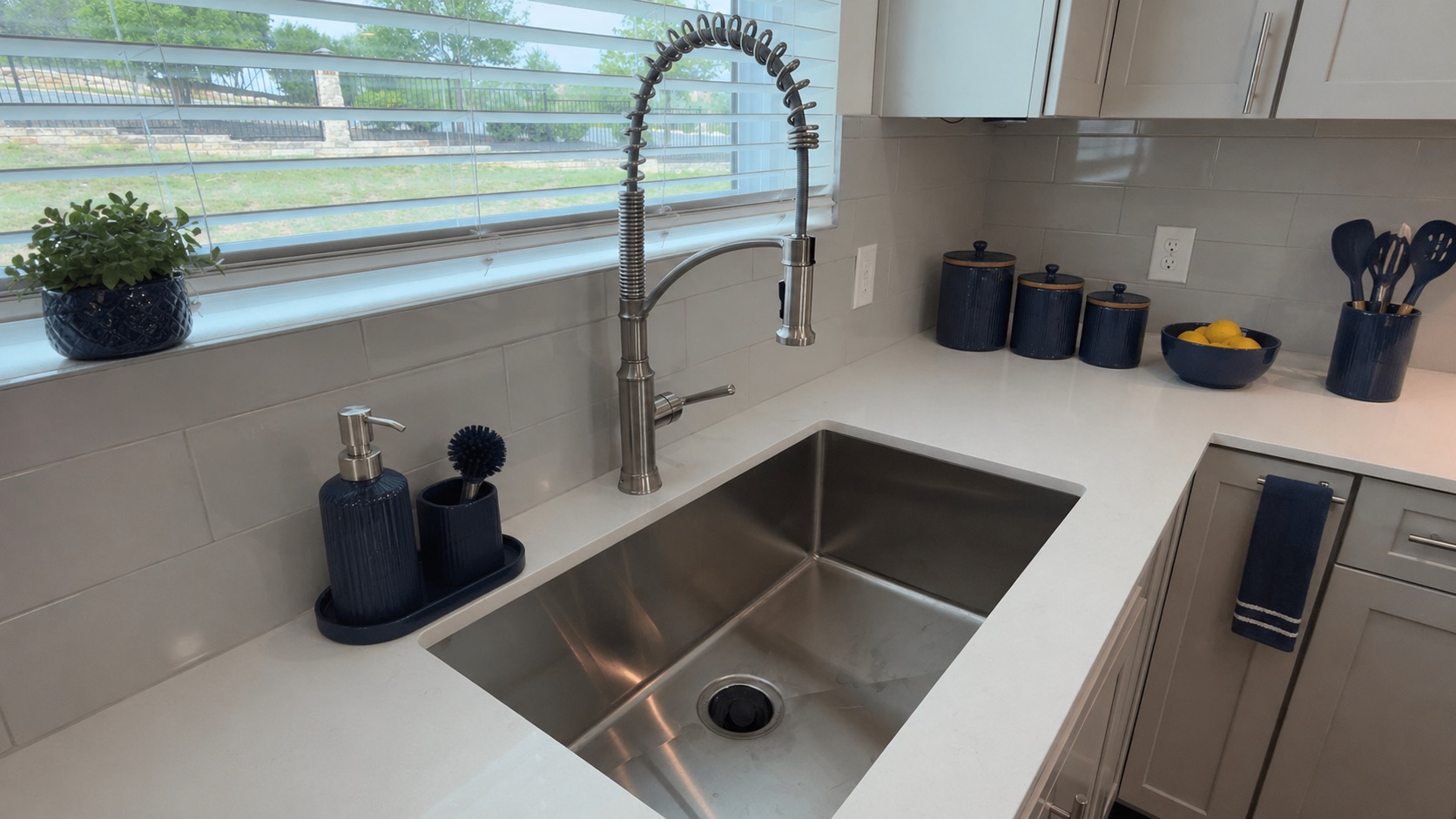 4300 Mansfield Dam Road, Unit 1021 Austin, TX 78734 - Photo 18 of 40 Kitchen sink area featuring a stainless steel undermount sink, a gooseneck faucet with a spring spout, light-colored Quartz countertops, a window with horizontal blinds, and a tiled backsplash