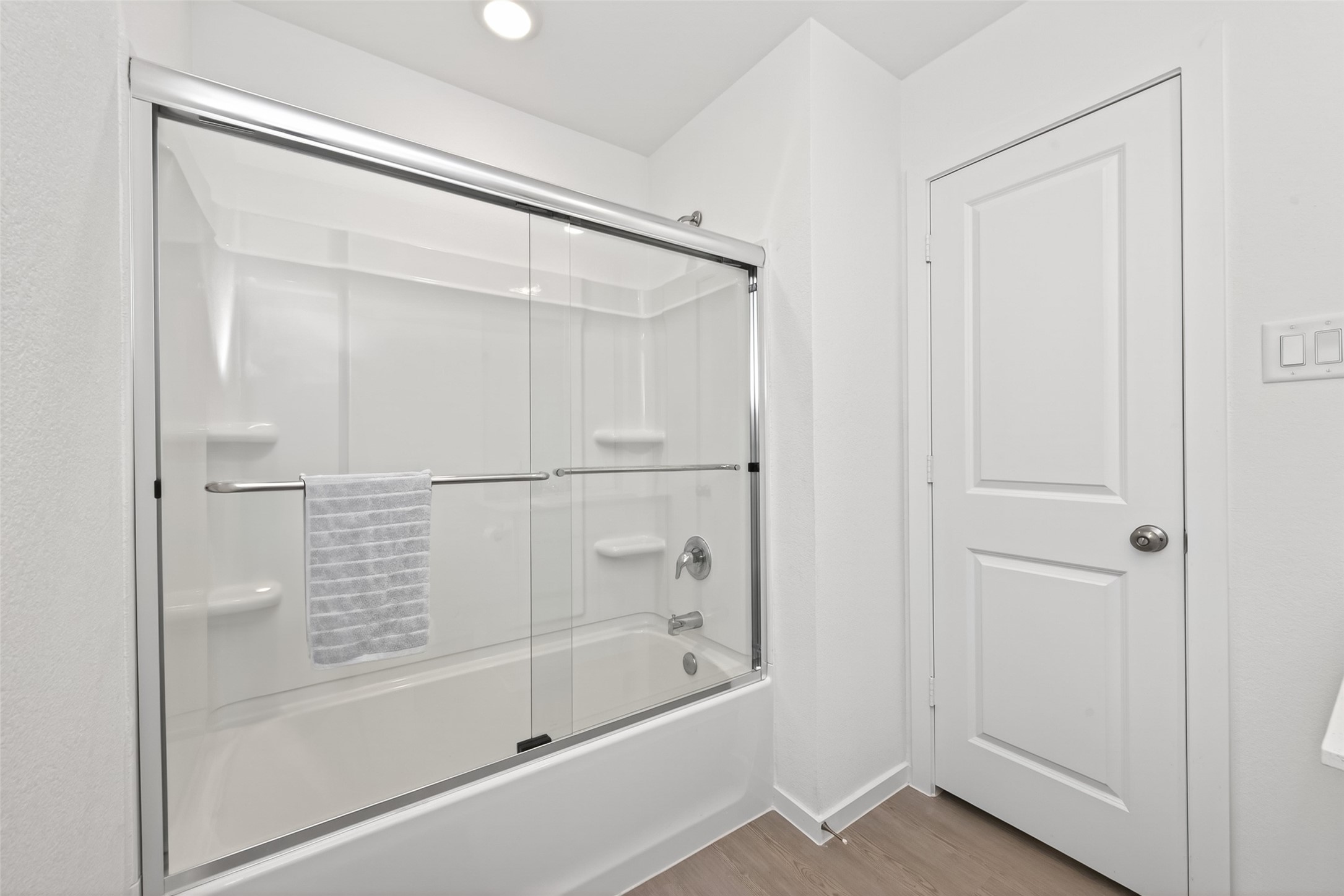 10720 Ellerston Road Cleveland, TX 77327 - Photo 20 of 32 This photo shows a modern, bright bathroom featuring a glass-enclosed bathtub with a shower, white walls, and a wooden floor. There is a white door and recessed lighting, creating a clean and fresh look.