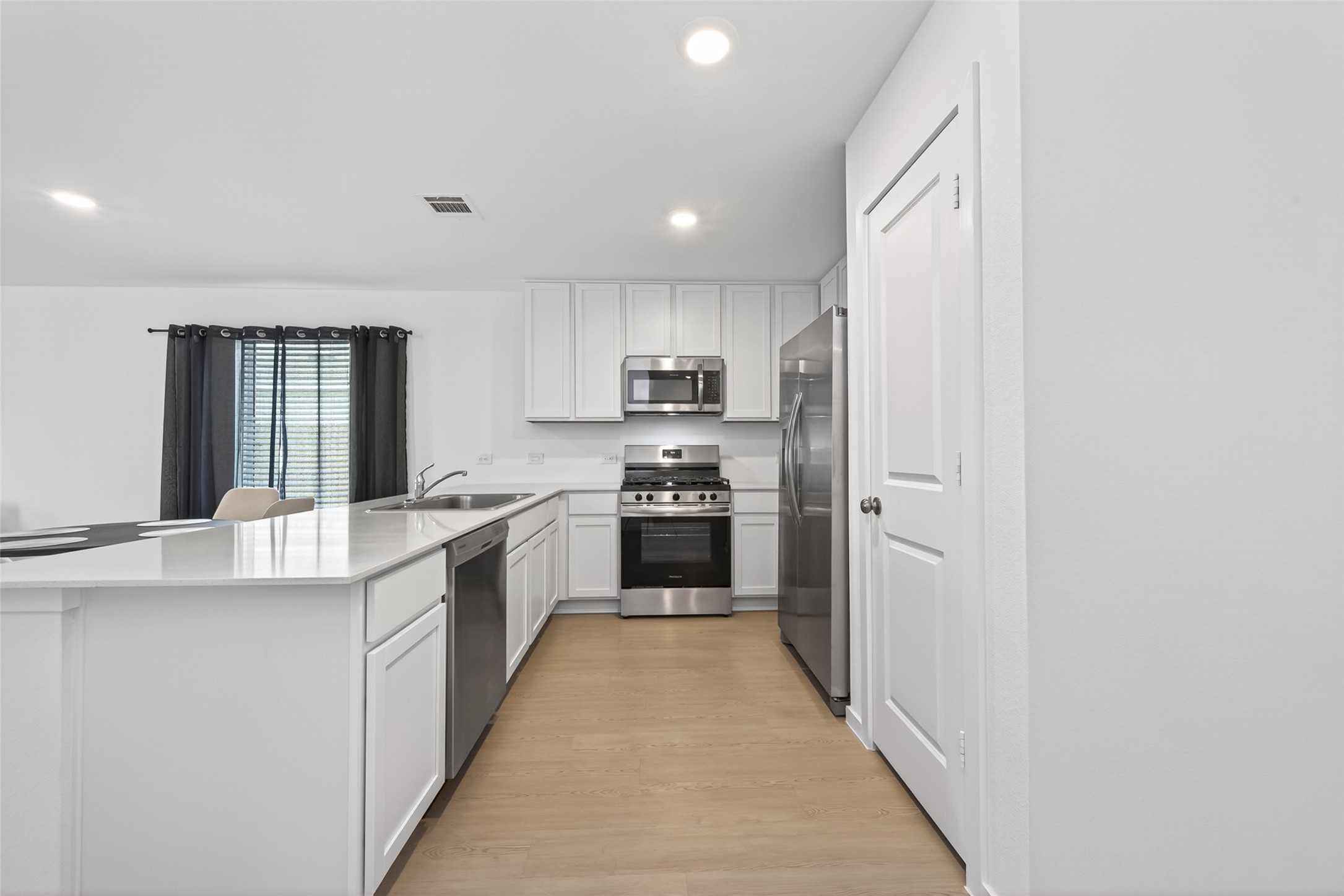 10720 Ellerston Road Cleveland, TX 77327 - Photo 4 of 32 This modern kitchen features sleek white cabinetry, stainless steel appliances, a spacious island with a sink, and natural light from a large window with dark curtains.