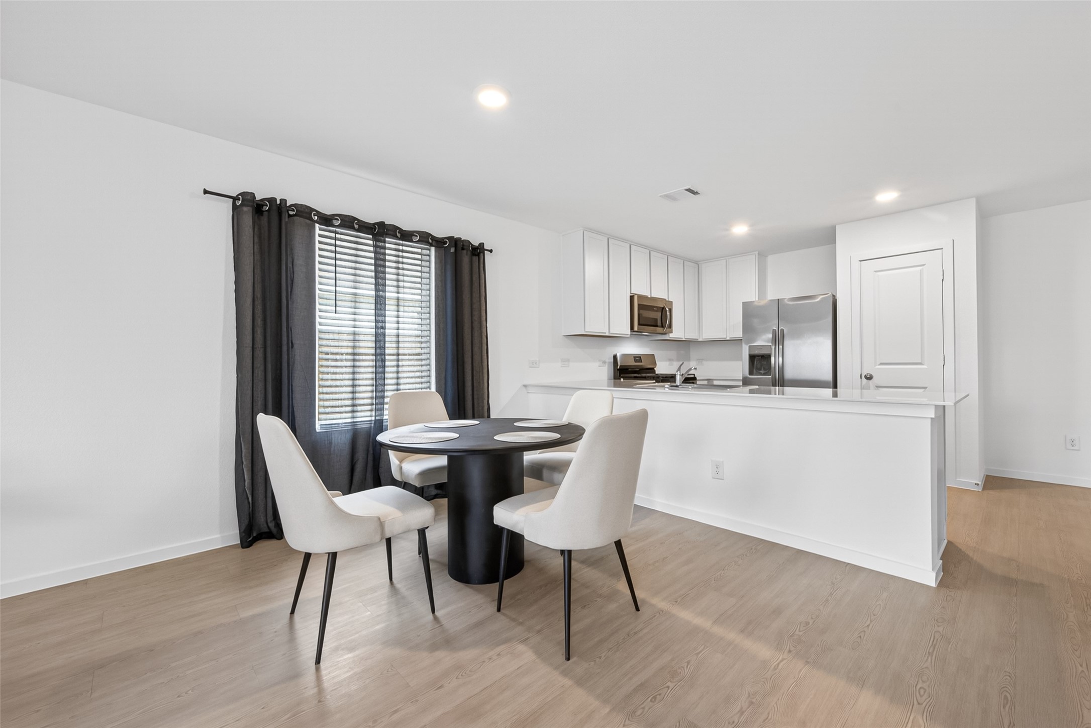 10720 Ellerston Road Cleveland, TX 77327 - Photo 9 of 32 Bright, modern kitchen and dining area featuring sleek white cabinetry, stainless steel appliances, and a cozy round dining table. Ample natural light and hardwood floors create a welcoming atmosphere.