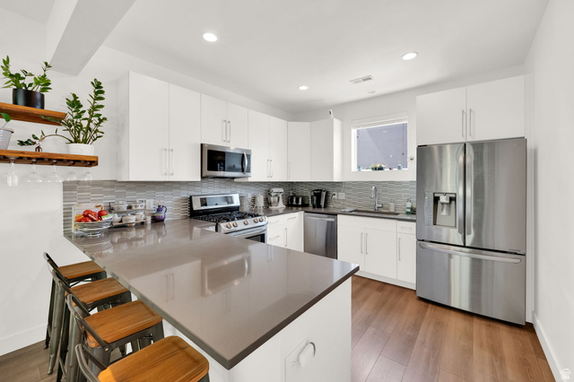 7713 Navarro View Court Midvale, UT 84047 - Photo 11 of 41 Kitchen featuring a peninsula with countertop seating, stainless steel appliances, wood-finish flooring, white cabinetry, and a subway tile backsplash