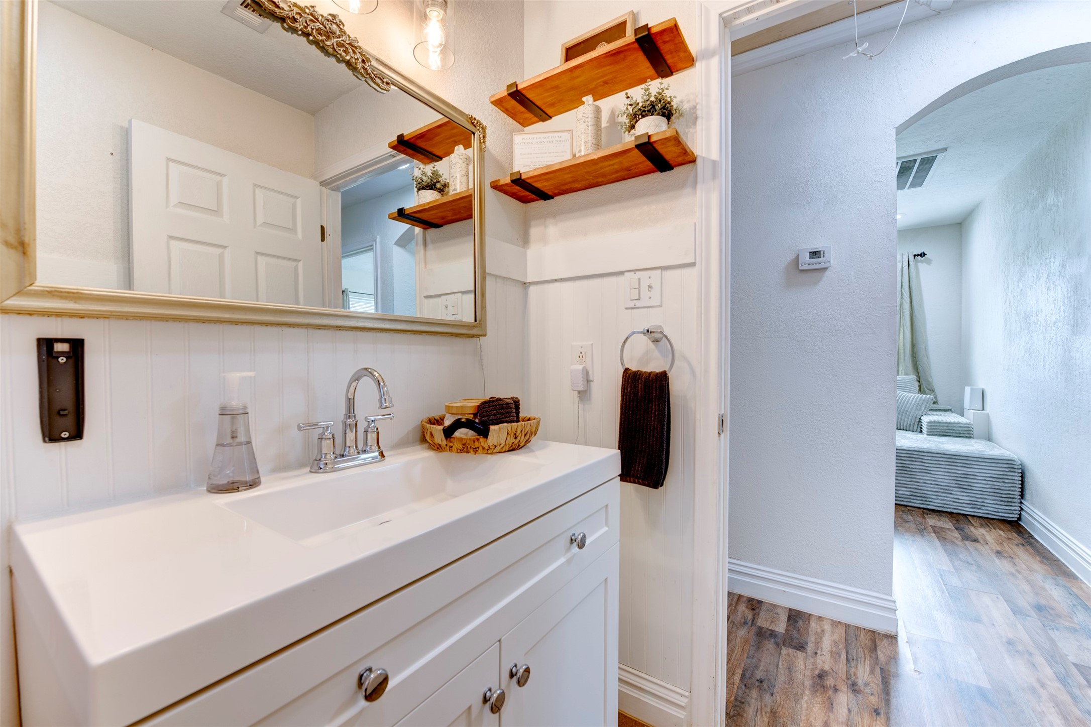 1121 Panama Street Houston, TX 77009 - Photo 21 of 35 Bathroom vanity close-up showing white shaker cabinetry with chrome knobs, single-basin white countertop, chrome bridge faucet, ornate silver-framed mirror, dual natural wood floating shelves with black iron brackets, beadboard wainscoting, and open arched hallway leading to bedroom.