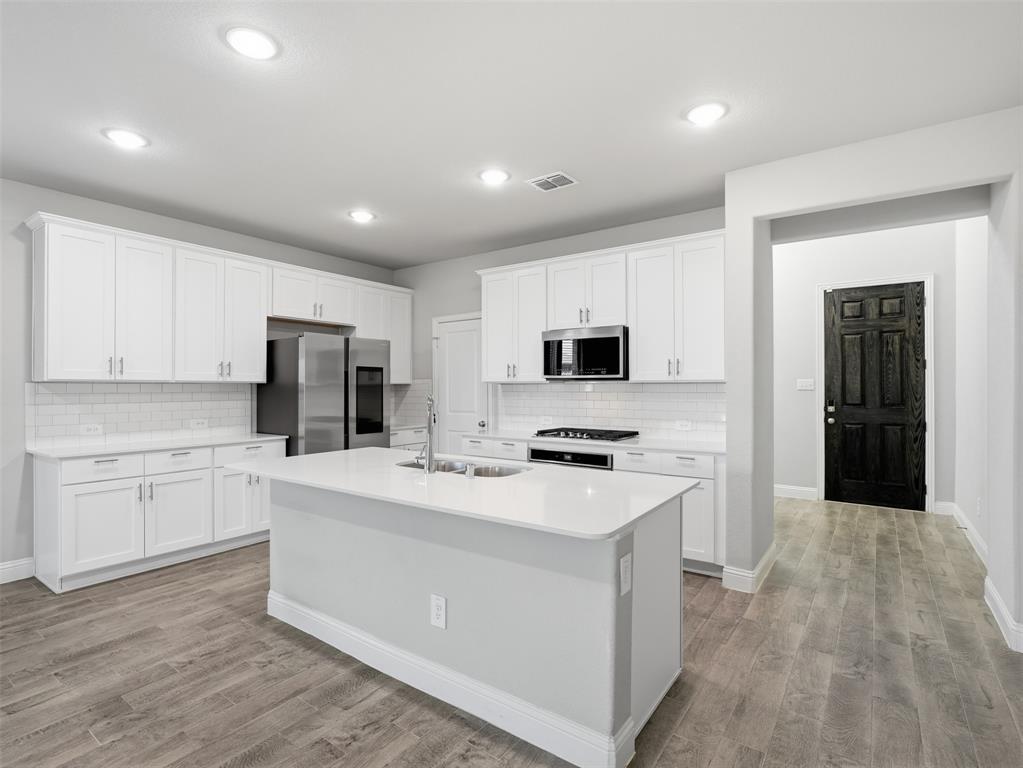 Kitchen featuring an island with an integrated sink, white cabinetry, stainless steel appliances, white subway tile backsplash, and wood-finish flooring