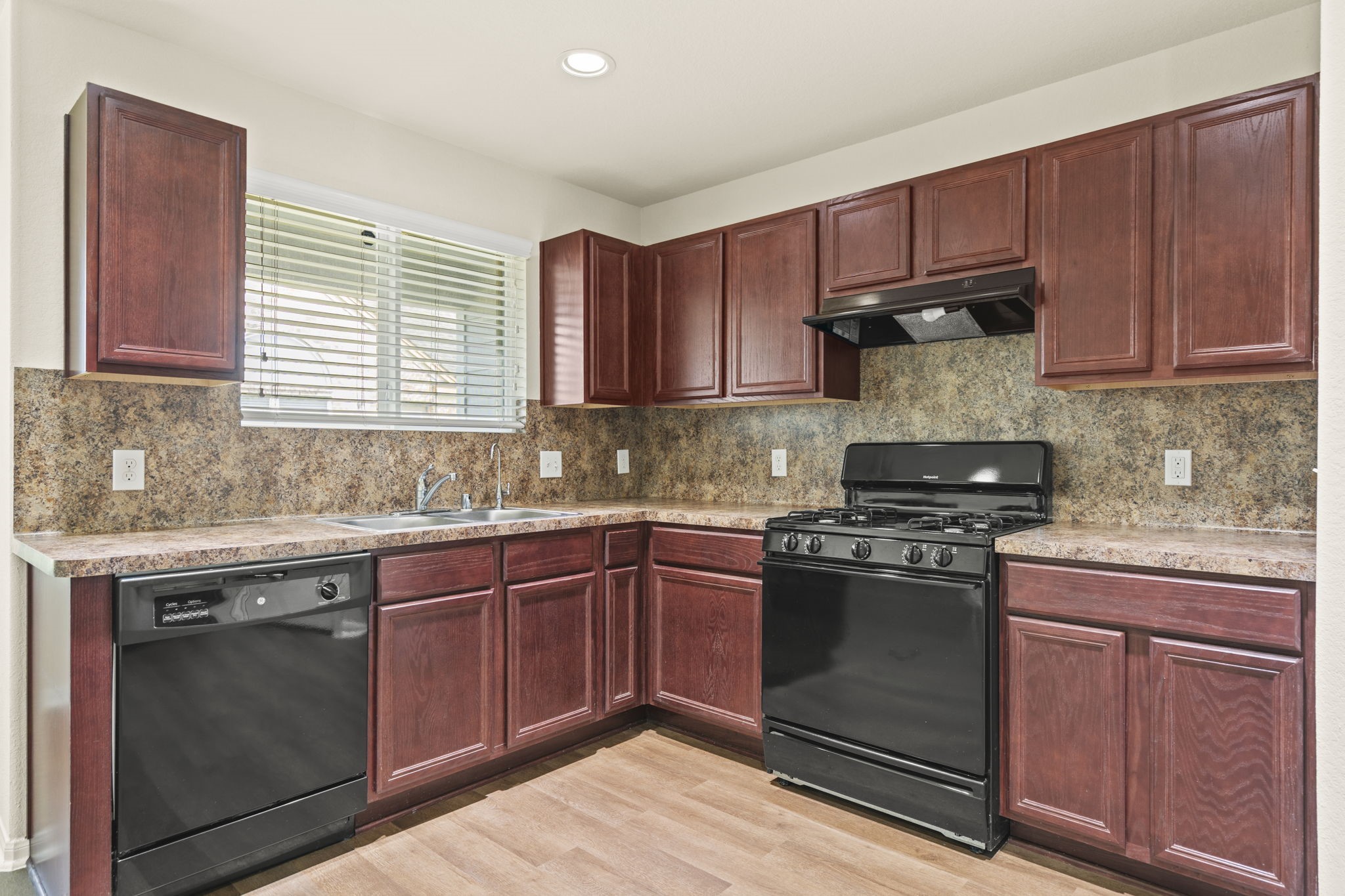 2110 Red Cedar Trail Rosenberg, TX 77471 - Photo 5 of 20 This close-up showcases an efficient L-shaped kitchen footprint complete with abundant wood-grain cabinetry and textured countertops with a matching integrated backsplash. Features include a double stainless steel sink under a window with fitted blinds, seamlessly integrated black appliances, and durable plank flooring.