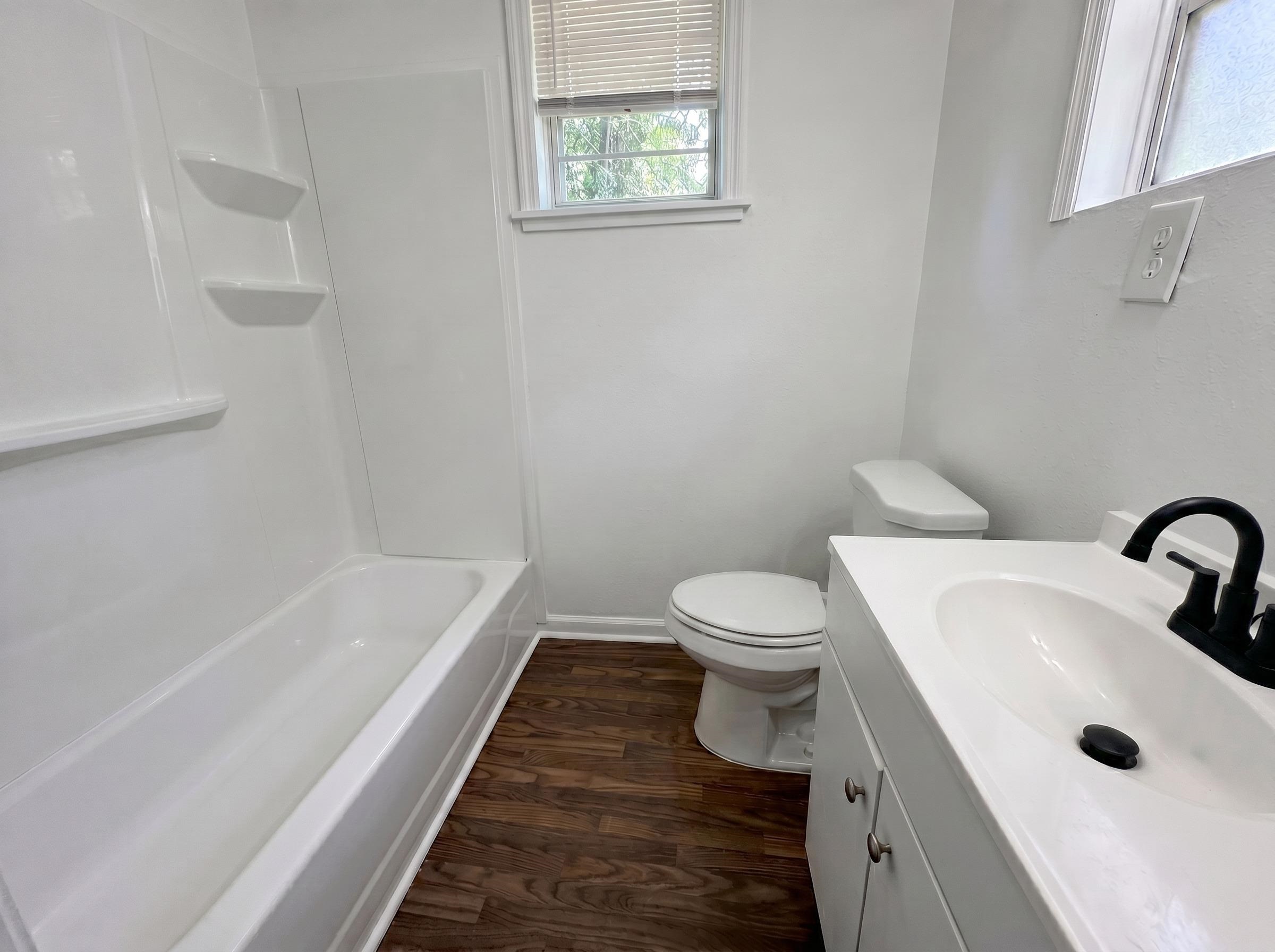 278 Hewlett Road Memphis, TN 38109 - Photo 5 of 5 Bathroom featuring white wall paint, wood-finish flooring, a white bathtub with integrated shelving, a white toilet, and a white vanity with a black faucet