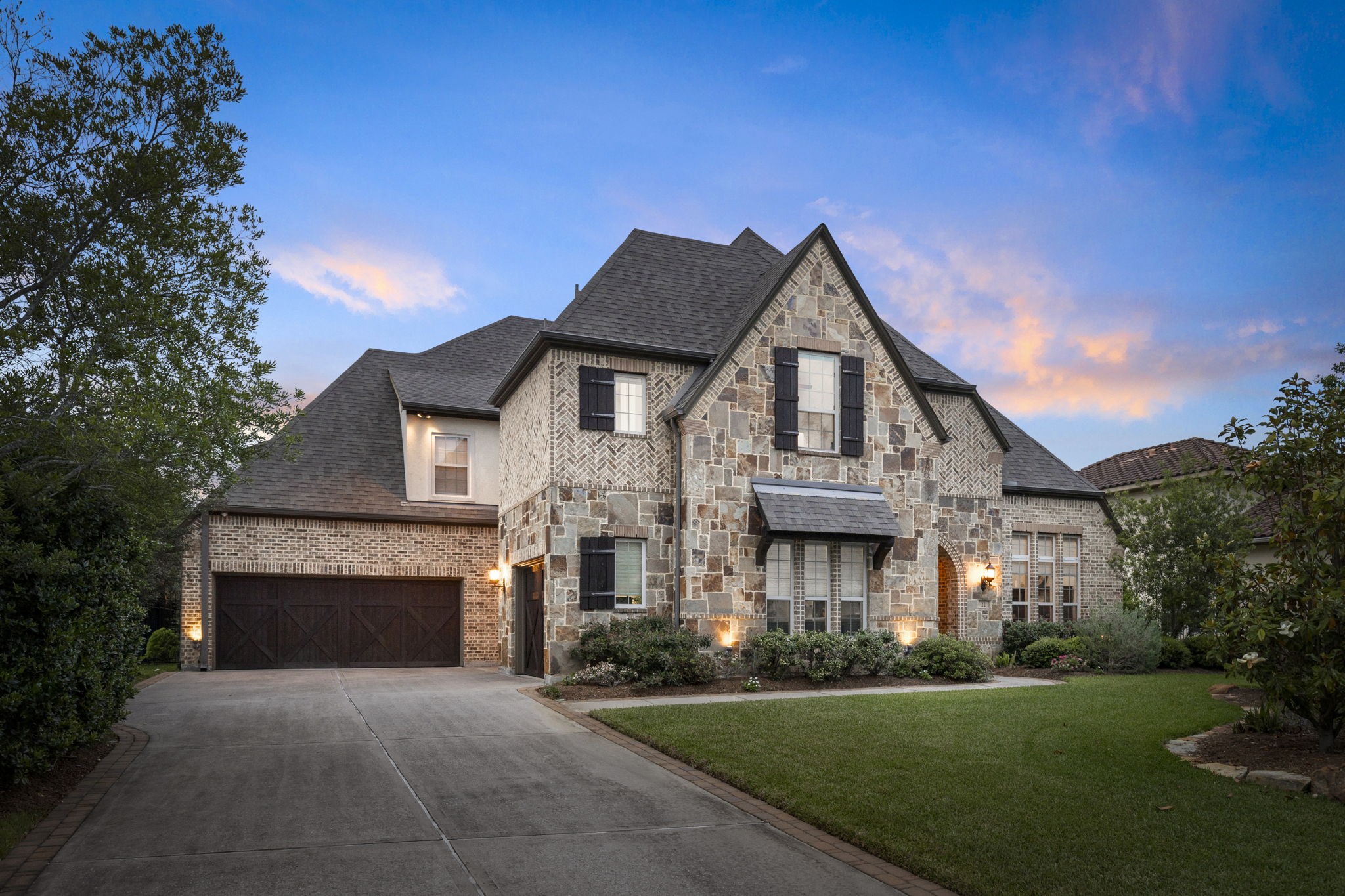 Elegant stone and brick façade with steep gables and charming shutters, beautifully illuminated at dusk. Wide drive with carriage-style garage doors enhances the home’s timeless curb​​‌​​​​‌​​‌‌​‌‌​​‌​​​​‌​​​‌‌​‌‌‌ appeal.
