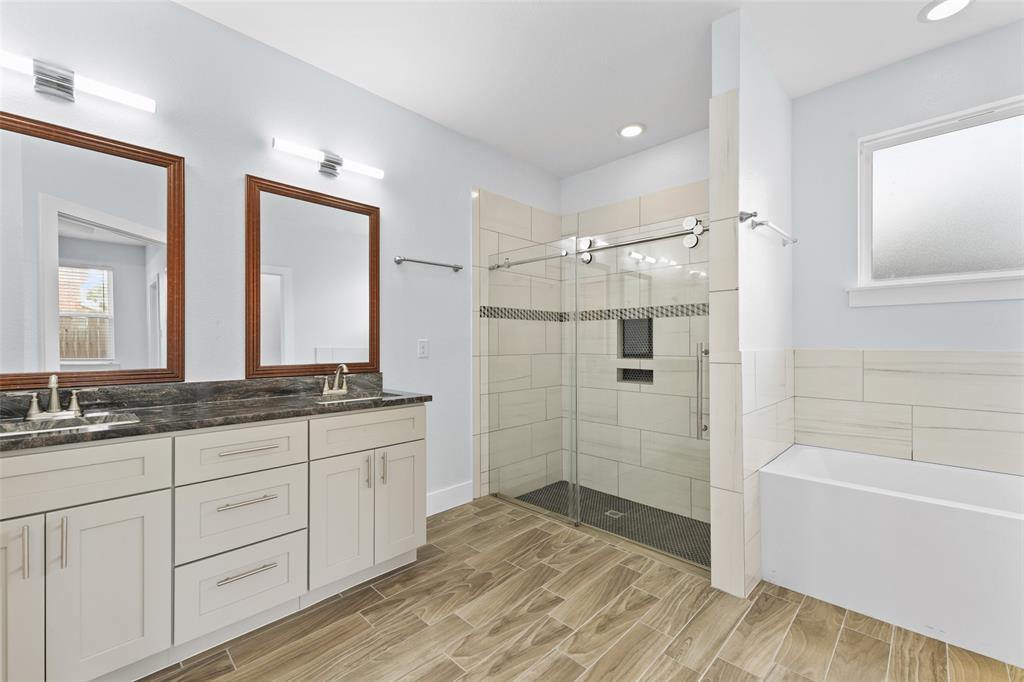 3807 Winding Road Arlington, TX 76016 - Photo 13 of 23 Spacious bathroom featuring a dual vanity with a dark countertop, wood-finish tile flooring, a glass-enclosed shower with a decorative tile band, and a built-in bathtub