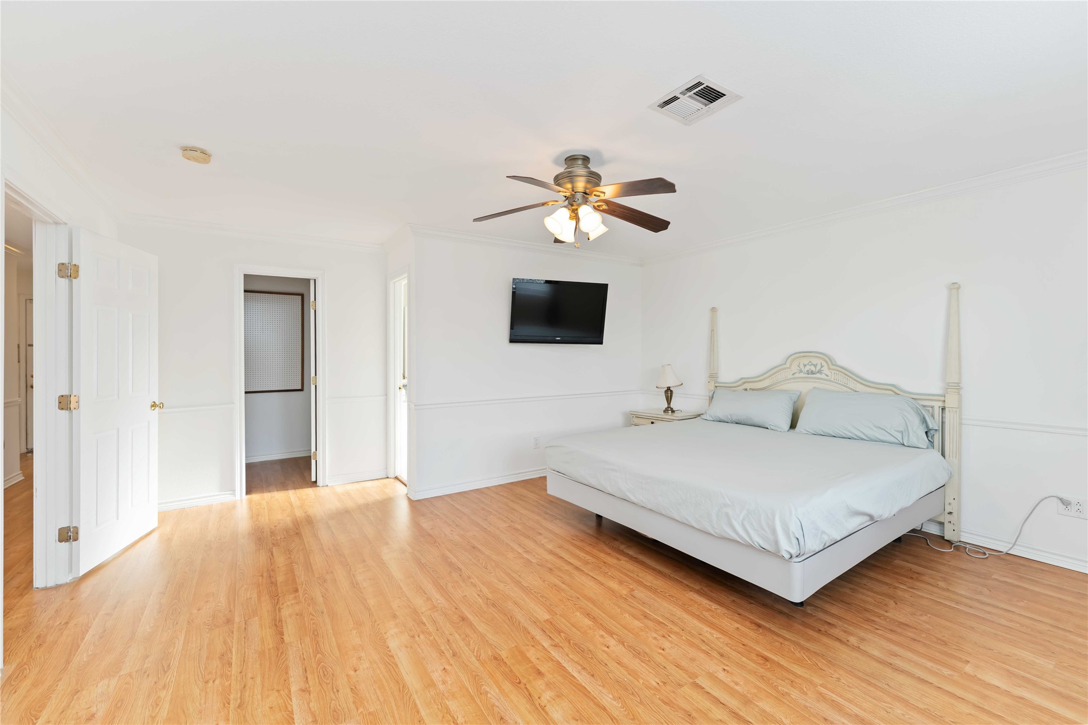 606 Packer Court Webster, TX 77598 - Photo 10 of 28 Spacious primary bedroom with light wood flooring, a ceiling fan, and ample natural light.
Features a large bed, wall-mounted TV, and access to an adjoining room. Clean, neutral decor creates a bright and welcoming atmosphere.