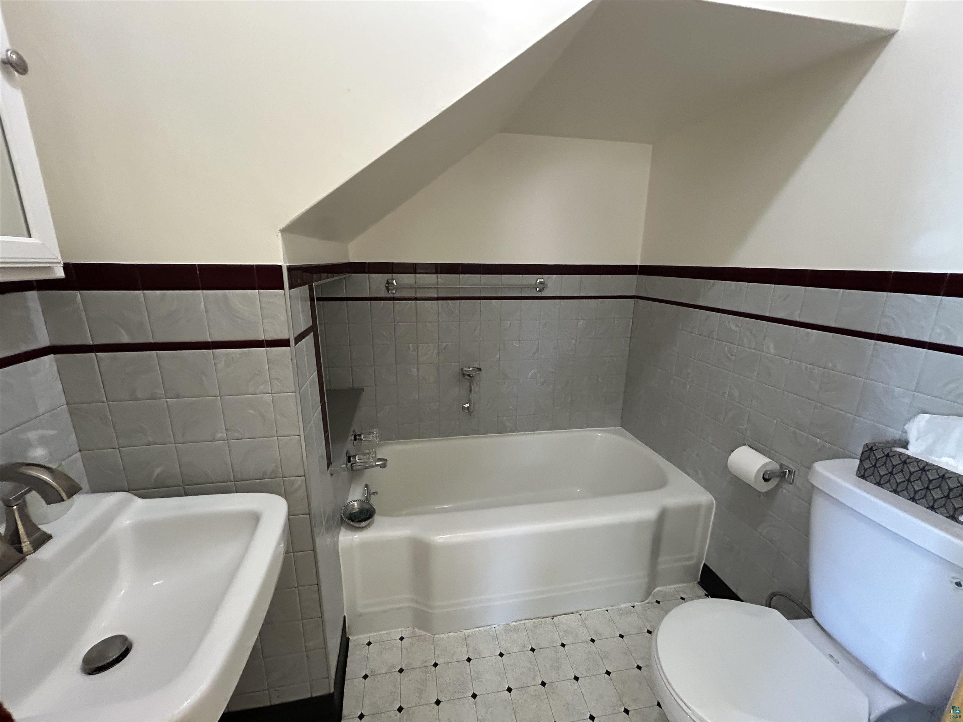 5090 London Road, Unit 1 Duluth, MN 55804 - Photo 23 of 54 Bathroom featuring a pedestal sink with brushed nickel faucet, a white bathtub, and a ceramic tile wainscoting with a dark accent stripe