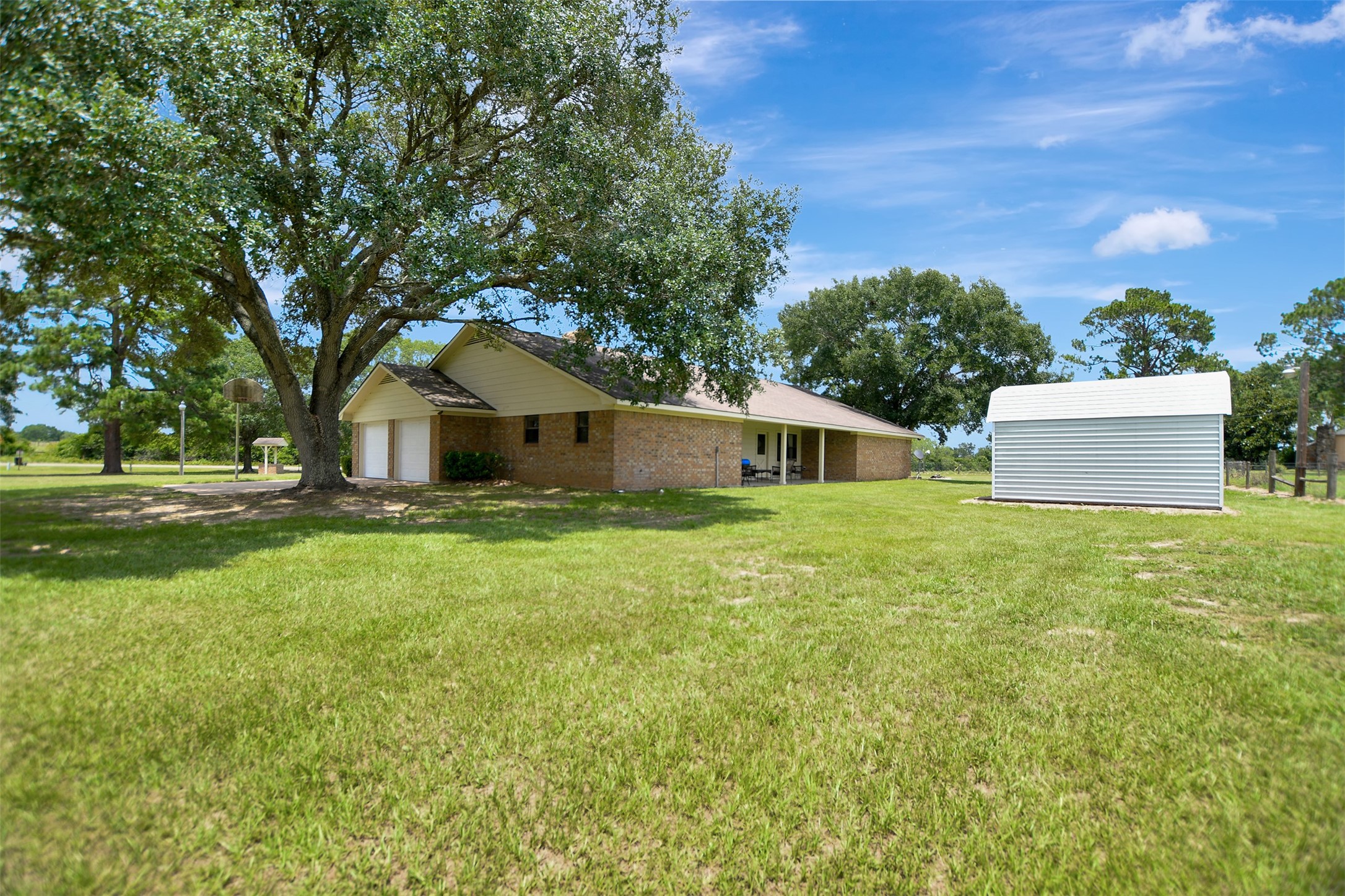 341 South S Loop Bellville, TX 77418 - Photo 29 of 33 This photo shows a spacious yard with a single-story brick home featuring a large tree providing shade. There's a covered patio and a separate white storage shed, offering ample outdoor space in a serene setting.