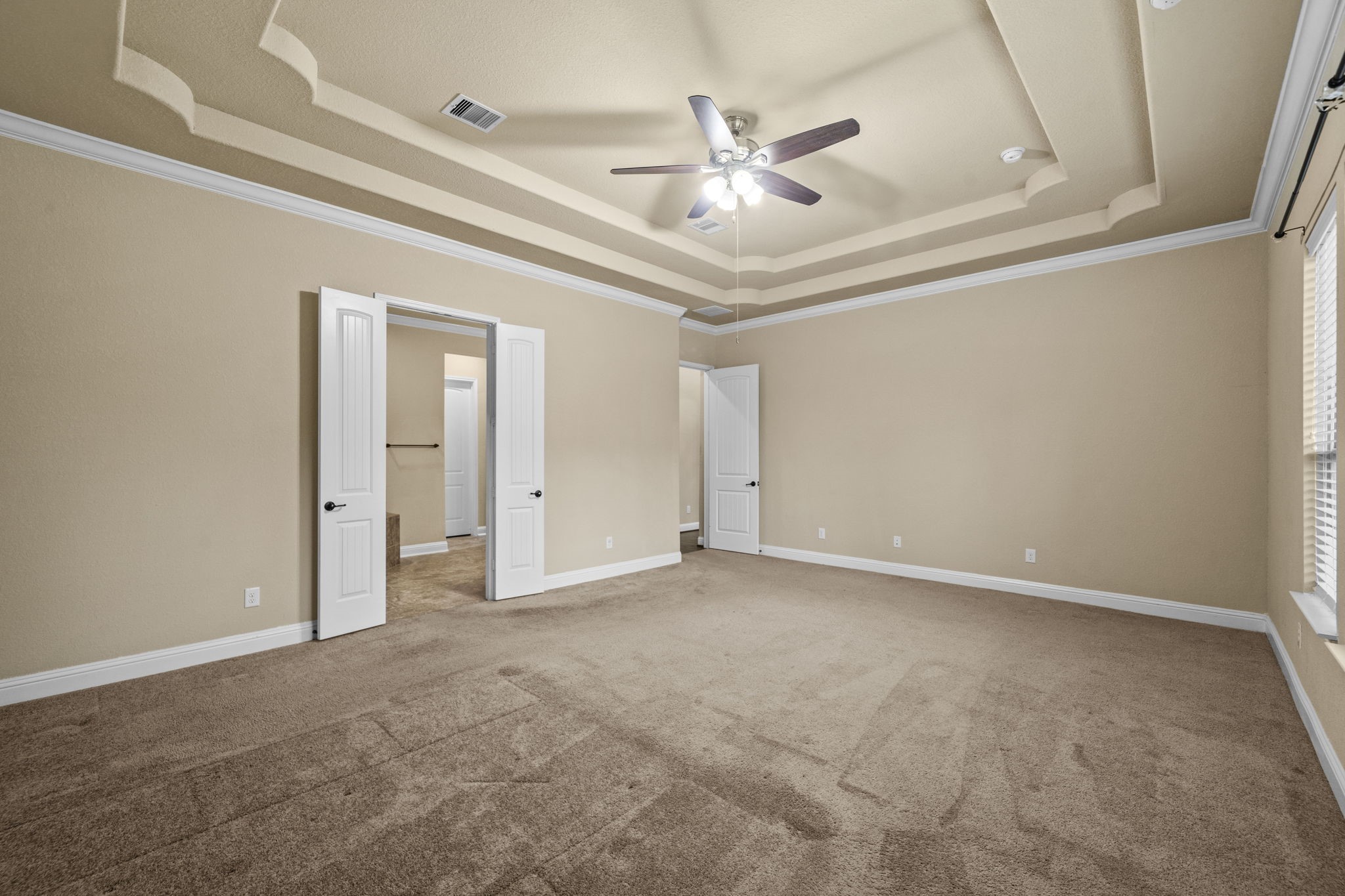 14215 Spindle Arbor Road Cypress, TX 77429 - Photo 31 of 47 This spacious bedroom features a tray ceiling with a ceiling fan, neutral beige walls, and carpet flooring. It has ample natural light from large windows and includes double doors leading to an en-suite bathroom or closet.