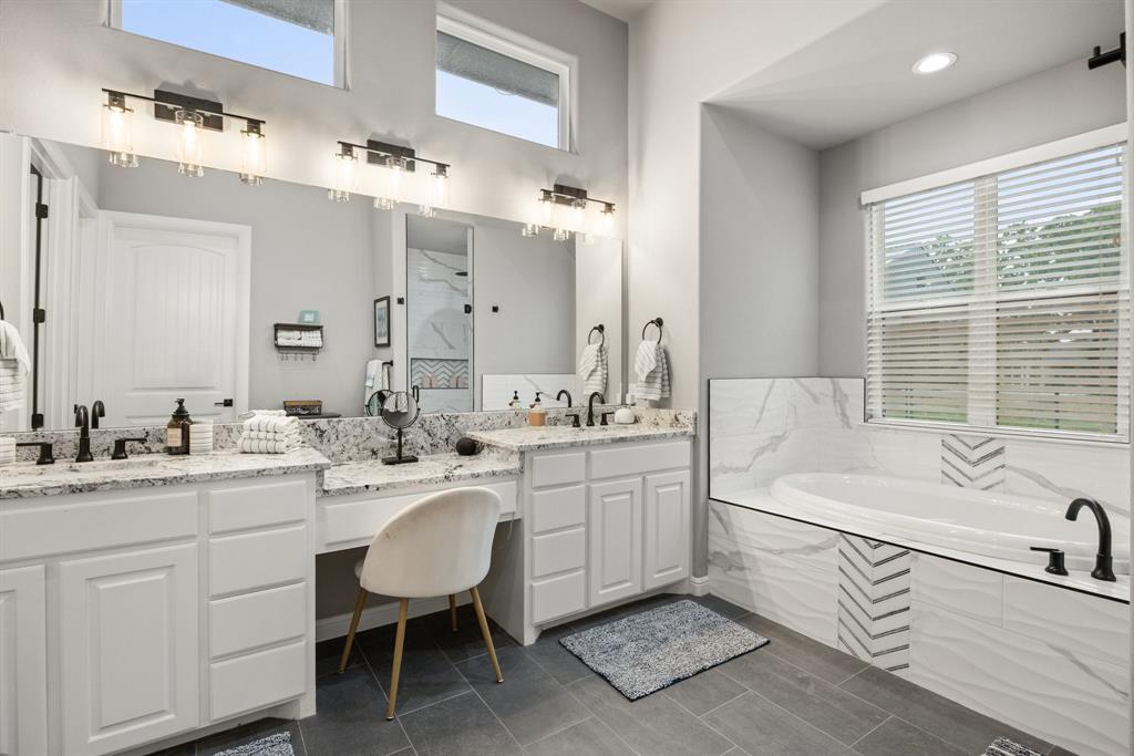 1800 Knox Road Tolar, TX 76476 - Photo 16 of 38 Spacious bathroom featuring dual granite-topped vanities, a built-in makeup area, a large soaking tub with marble-look tile surround, and contemporary dark bronze fixtures