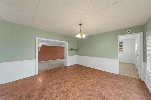 an empty room with chandelier fan and kitchen view