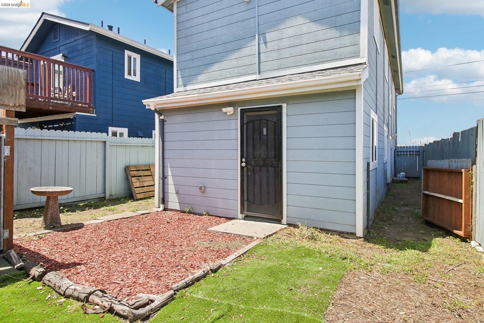 874 Pine Street Oakland, CA 94607 - Photo 26 of 39 Light blue siding exterior featuring a black entry door with security screen, a side walkway, a fenced perimeter, and a mulched patio area