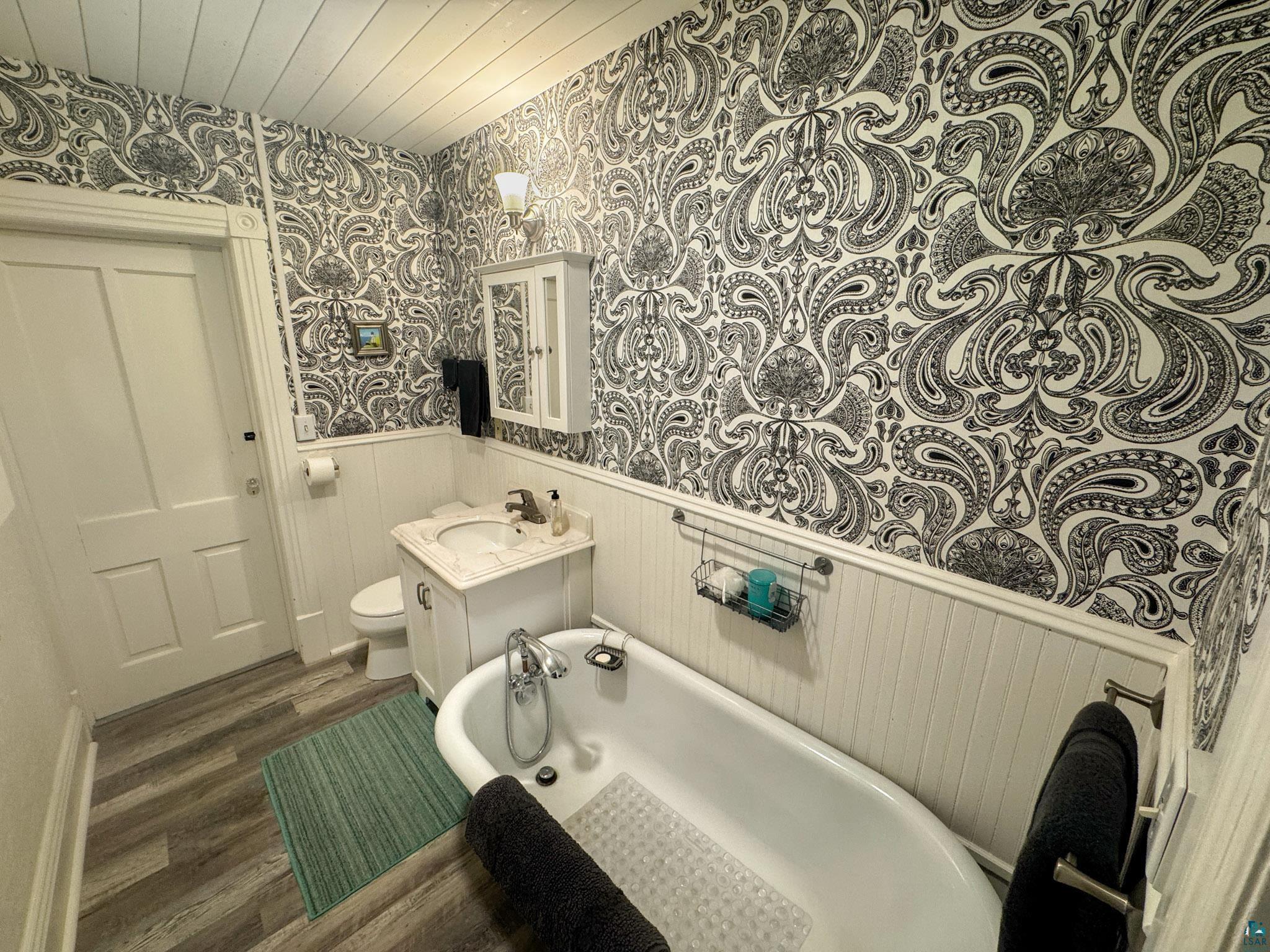 303 East 5th Street Washburn, WI 54891 - Photo 31 of 34 Bathroom featuring a clawfoot bathtub, single vanity with a decorative stone countertop, black and white patterned wallpaper, white beadboard wainscoting, and wood-finish flooring