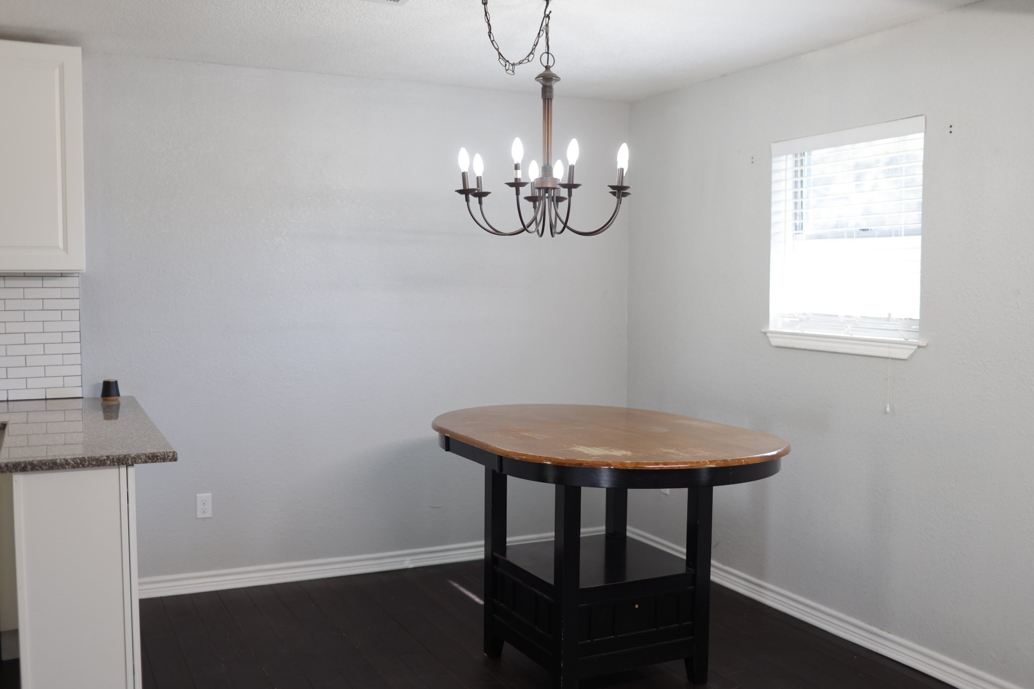 407 Tamarind Street Lake Jackson, TX 77566 - Photo 10 of 36 This photo shows a dining area with a simple chandelier and a wooden table. It features a window for natural light, dark flooring, and is adjacent to a kitchen counter with a white tile backsplash.