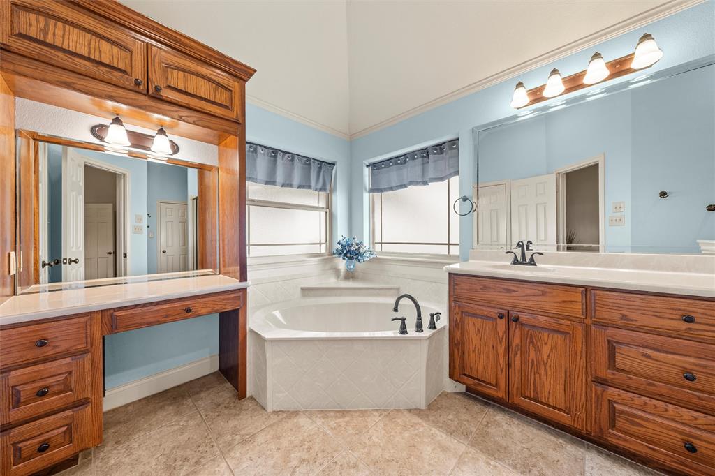 231 Trlica Road West, TX 76691 - Photo 21 of 36 This original primary bathroom offers a spacious, well-appointed layout with classic finishes and abundant storage. A large corner soaking tub is surrounded by windows that bring in natural light while maintaining privacy, creating a bright, relaxing atmosphere. Dual oak vanities provide generous counter space and cabinetry, while a built-in makeup vanity adds convenience and charm. Neutral tile flooring and elevated ceilings enhance the open feel, making this a functional and inviting primary bath.