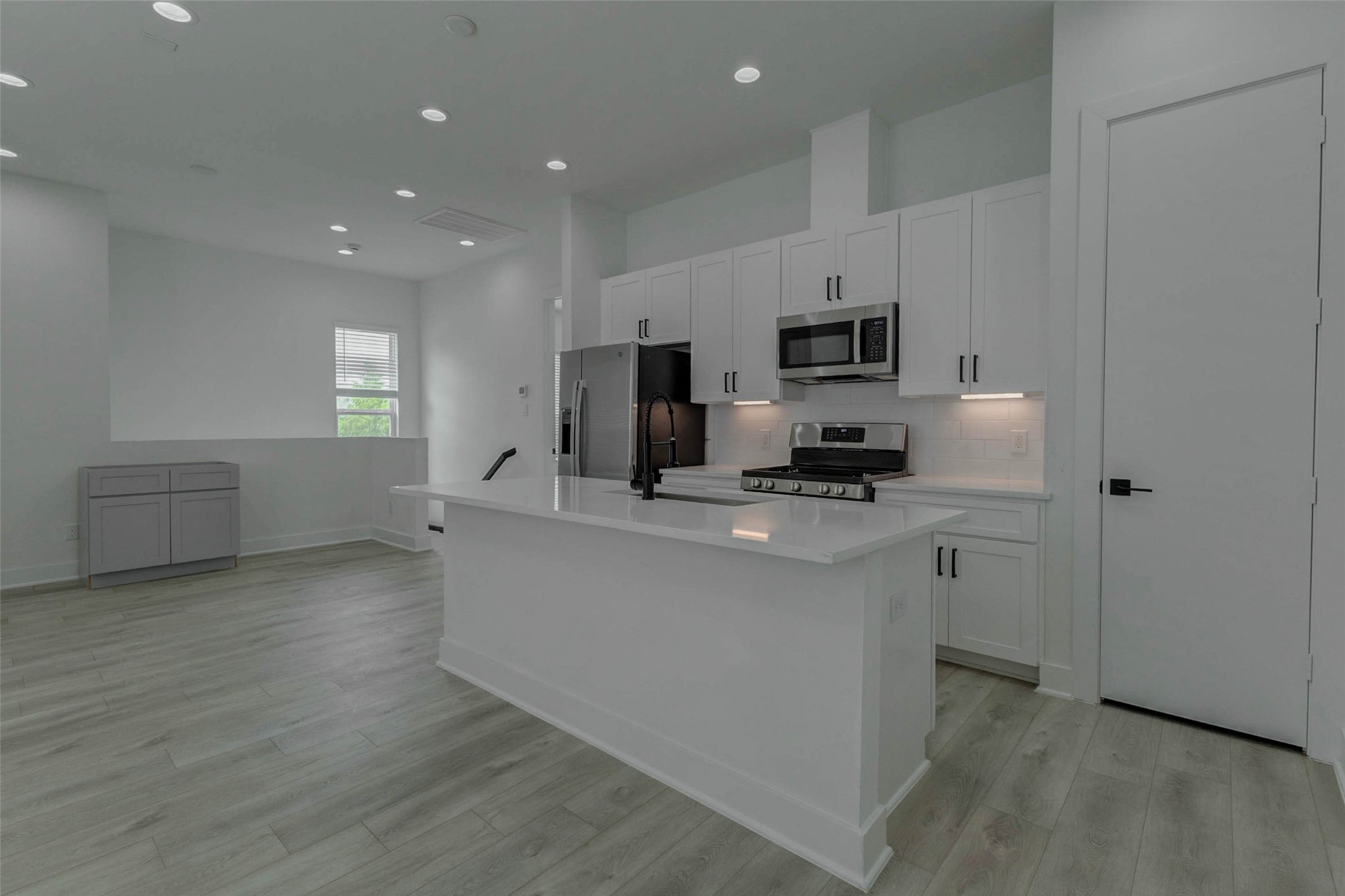5705 Elysium Street Houston, TX 77091 - Photo 14 of 22 Modern kitchen view featuring a large white island, quartz-style countertops, stainless appliances, gas range, built-in microwave, white shaker-style cabinetry, black hardware, and under-cabinet lighting. The open layout flows into the living area, creating a bright, contemporary space for cooking, dining, entertaining, and everyday living.