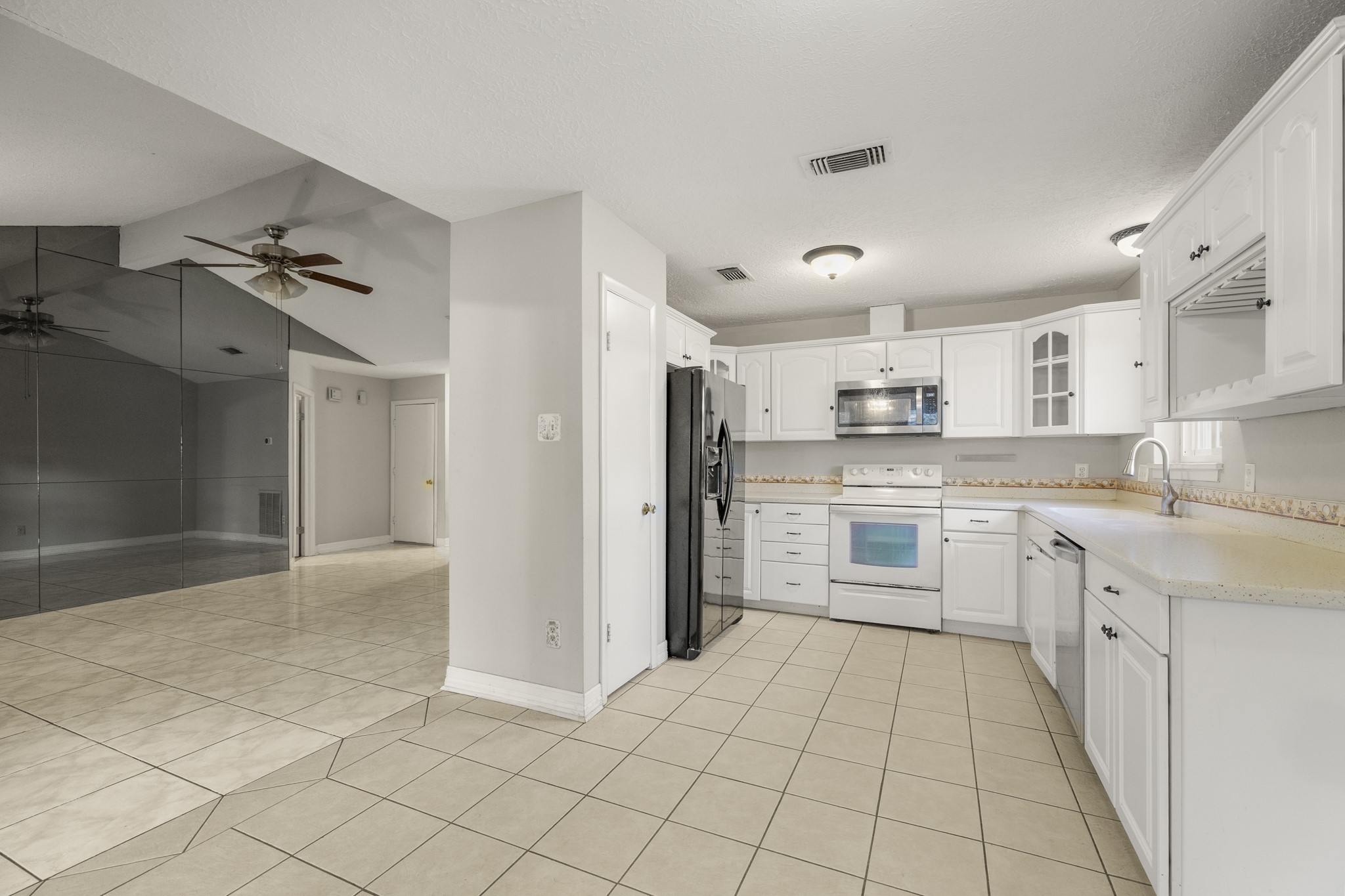 23826 Lestergate Drive Spring, TX 77373 - Photo 12 of 45 Turning from the living room, the kitchen comes into view just steps away, keeping conversation and everyday routines connected between the main gathering spaces. White cabinetry wraps the room with multiple banks of drawers for easy-access storage, while the range, microwave, refrigerator, and dishwasher are all positioned within a compact working area. Tile flooring continues underfoot, with the mirrored living room wall still visible to the left for a clear sense of where we are in the home.