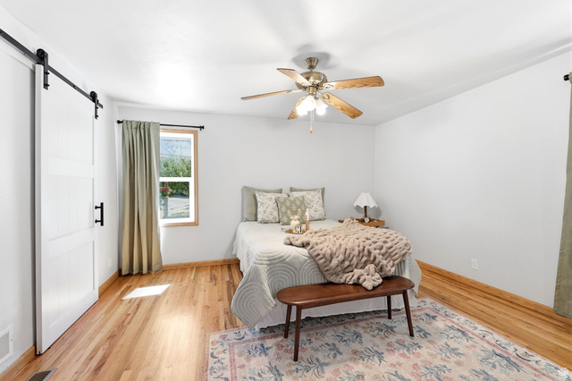 1180 North 400 East Bountiful, UT 84010 - Photo 22 of 50 Bedroom featuring wood-finish flooring, a ceiling fan with light fixture, a window with wood trim, and a white barn door with black hardware