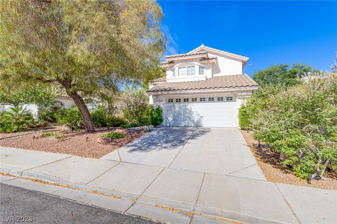 253 Camelback Ridge Avenue Henderson, NV 89012 - Photo 2 of 26 Mature trees frame a concrete driveway leading to a two-story home with tile roofing.