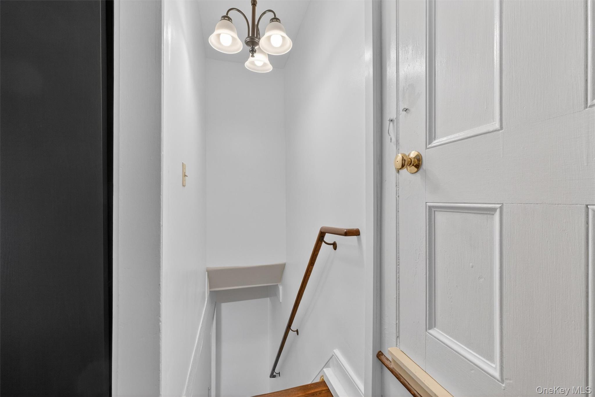 121 Old Glenham Road Fishkill, NY 12524 - Photo 34 of 50 Stairwell featuring a traditional three-light chandelier, a wood-finish handrail, white wall paneling, and a white paneled door with a brass knob connecting to a side bedroom from the dining room side staircase.