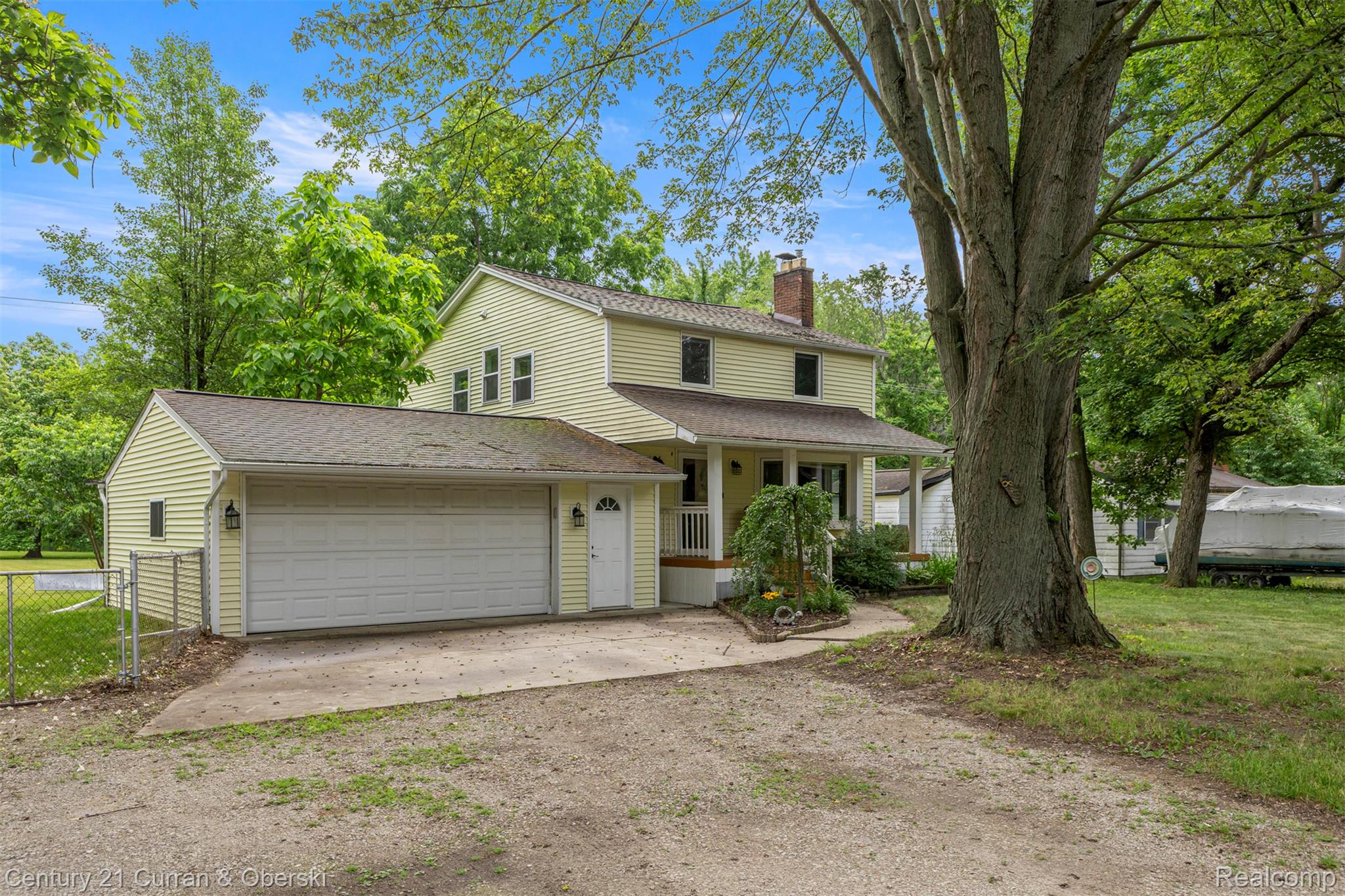 18491 Savage Road Belleville, MI 48111 - Photo 47 of 49 Traditional two-story residence featuring light yellow siding, an attached two-car garage with a white sectional door, a covered front porch, a prominent red brick chimney, and mature trees framing the property