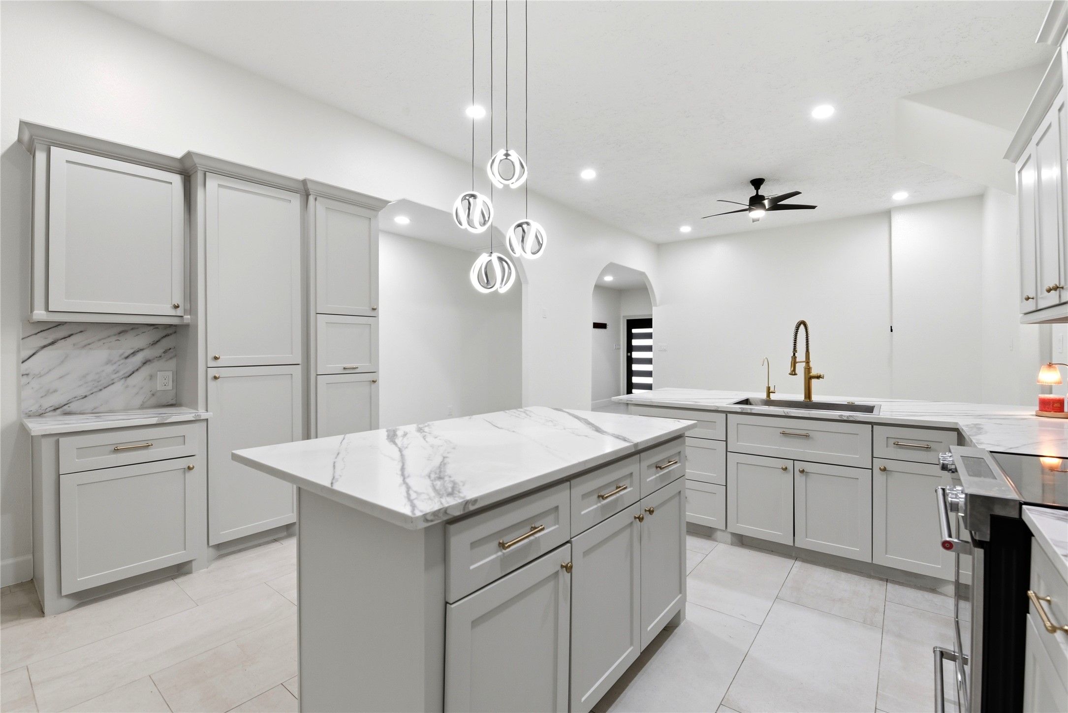 9403 Spaulding Street Houston, TX 77016 - Photo 15 of 50 This modern kitchen features sleek white cabinetry with gold hardware, marble countertops, and a large island. It includes contemporary lighting fixtures, stainless steel appliances, and a stylish ceiling fan, creating a bright and elegant space.