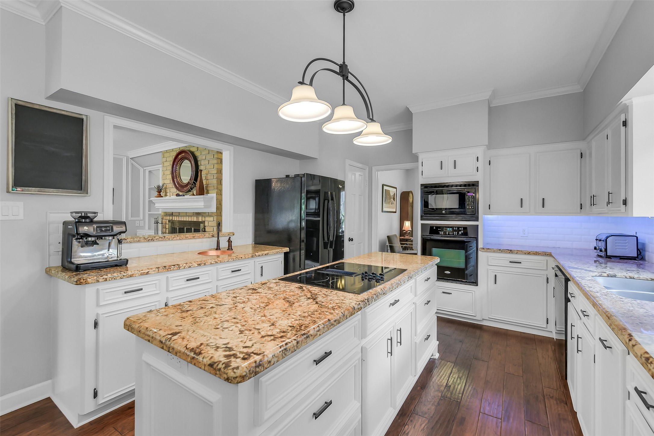 26 Split Rock Road Spring, TX 77381 - Photo 15 of 40 This kitchen features elegant granite countertops, white cabinetry, and modern appliances. It has a central island with a built-in cooktop, hardwood floors, and a cozy view into a living area with a fireplace.