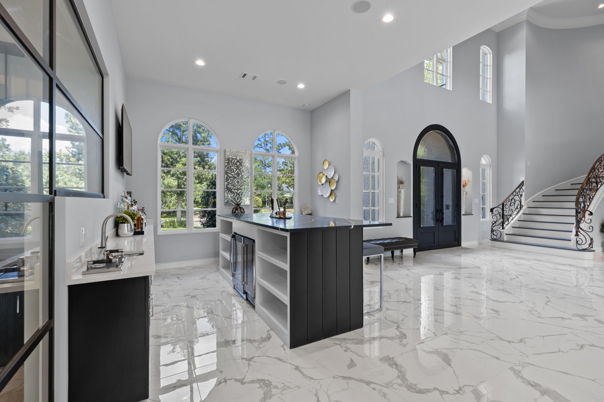 39 Maymont Way Spring, TX 77382 - Photo 9 of 50 This spacious, elegant foyer features high ceilings, large arched windows, and a stunning curved staircase with ornate railings. The area is bright with natural light, complemented by glossy marble floors and a sleek, modern bar area.
