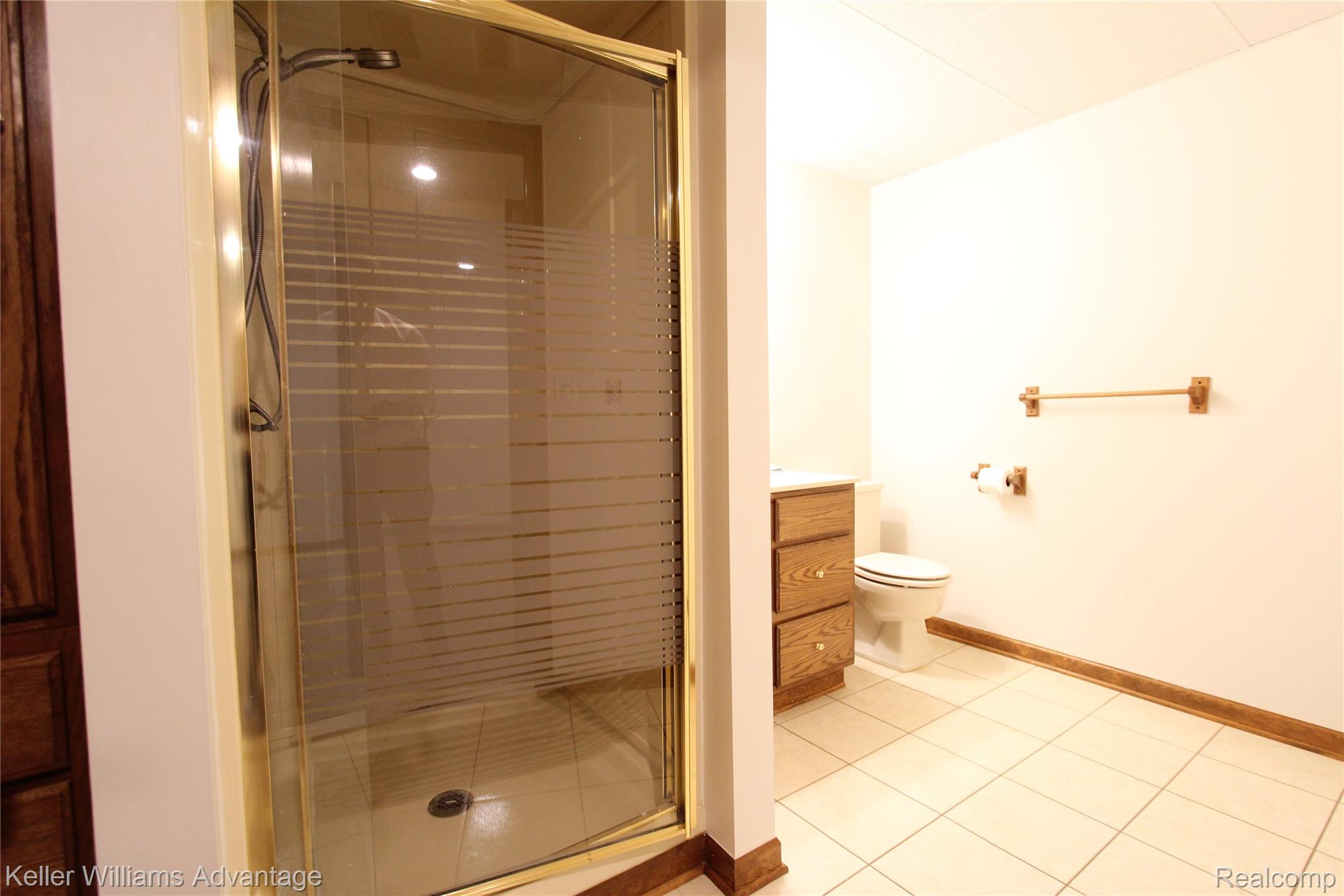 21850 River Ridge Trail Farmington, MI 48335 - Photo 24 of 33 Bathroom featuring a shower enclosure with gold-tone framing and frosted glass, light-toned ceramic tile flooring, a white vanity with wood cabinetry, a white toilet, and wood baseboards