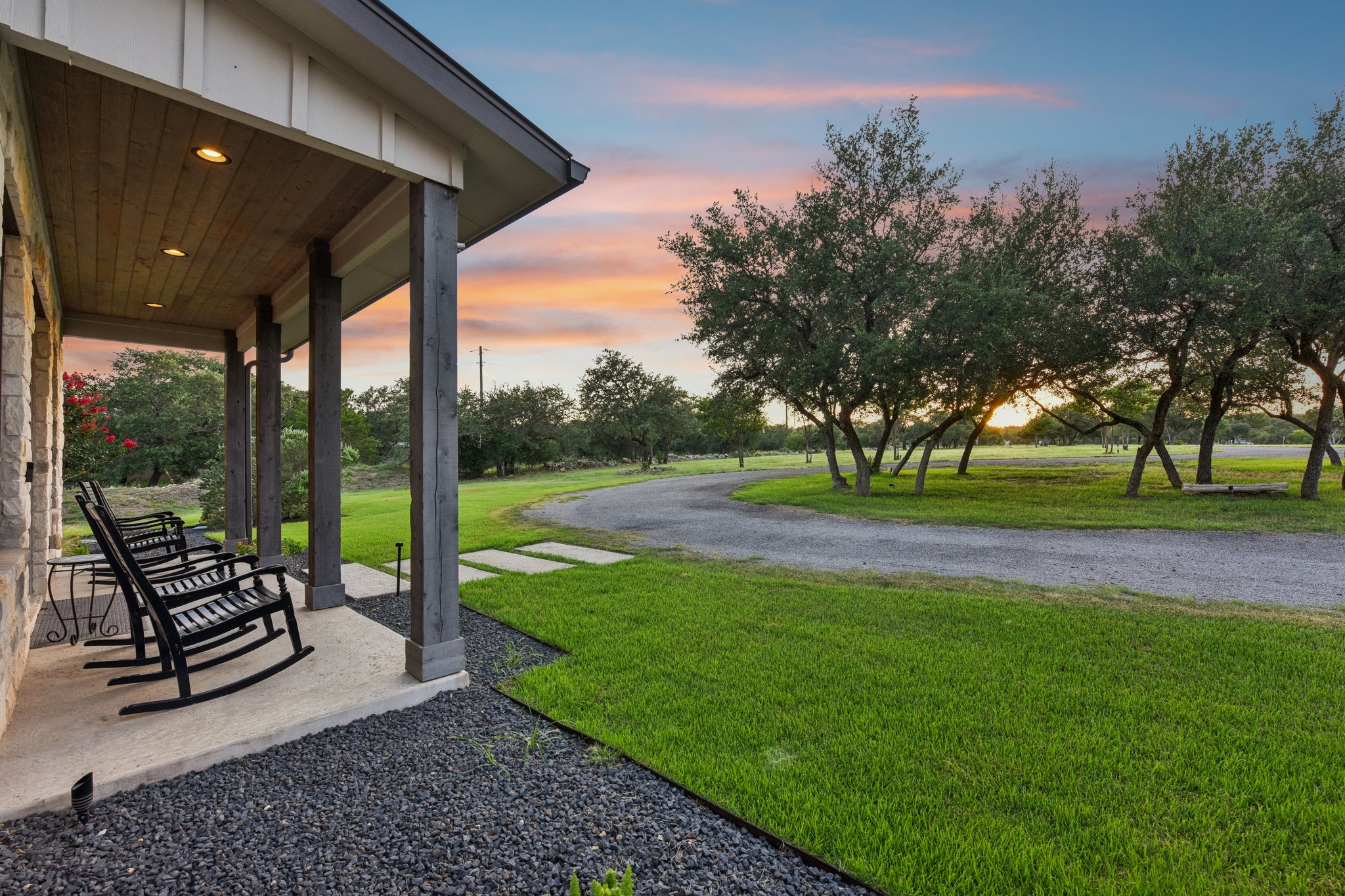 2203 Improver Road Spicewood, TX 78669 - Photo 11 of 38 An inviting front porch with dark rockers overlooks a sprawling lawn and winding driveway, framed by mature trees under a pastel sunset.