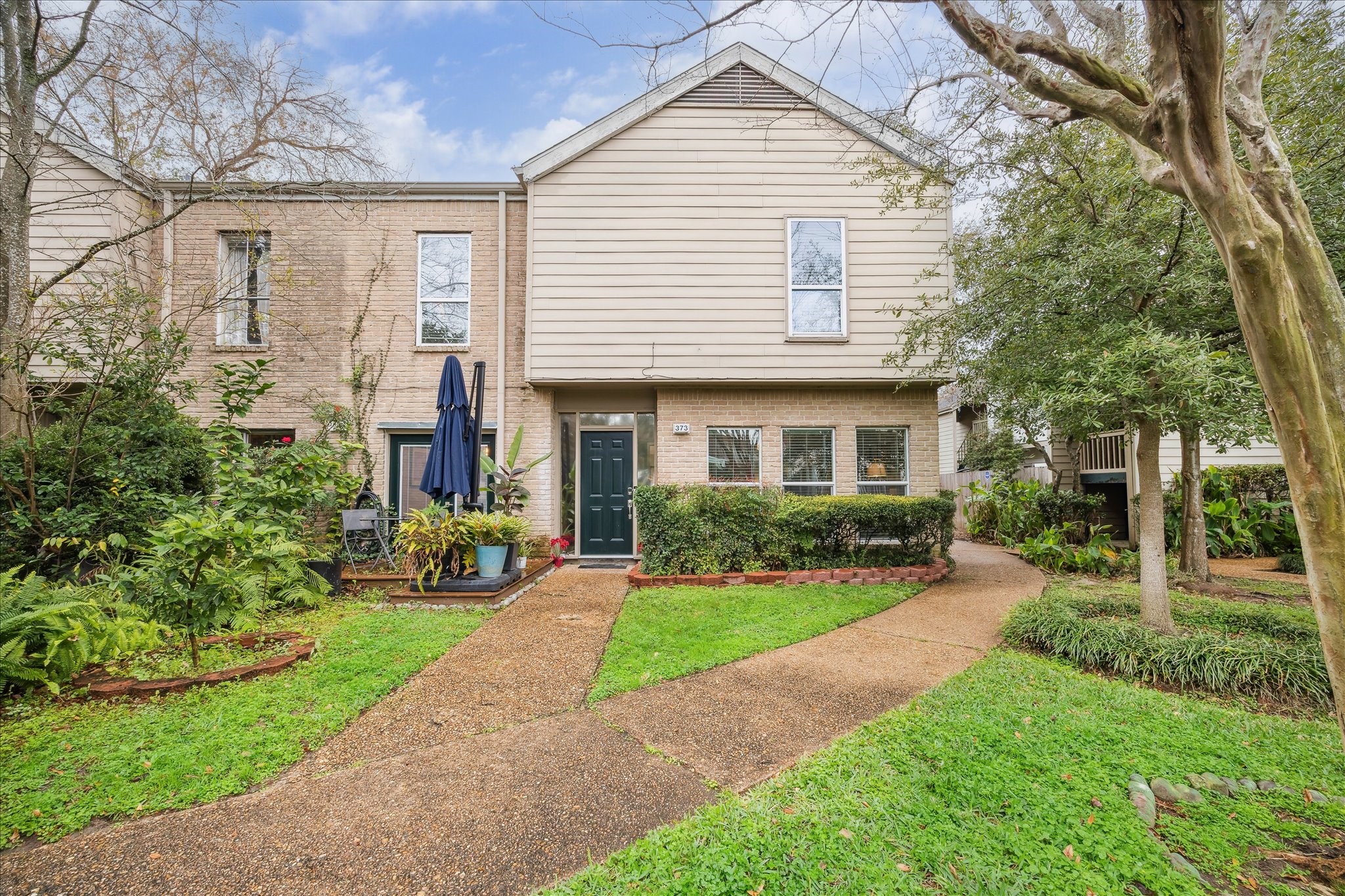 Charming two-story 3/2/1 townhouse with a welcoming front garden and pathway. Features a mix of brick and siding exterior, with a cozy front patio area. Lush greenery surrounds the home, creating a serene atmosphere.
