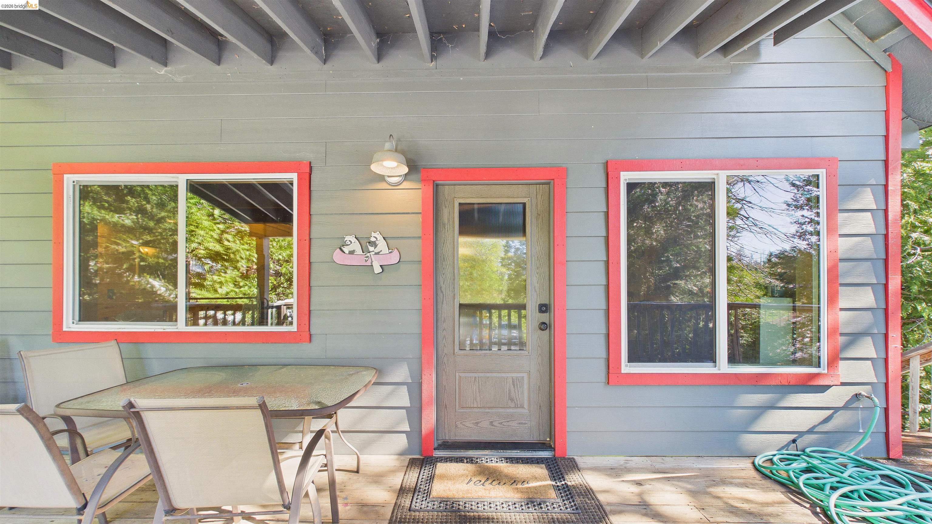 26570 Crystal Road Long Barn, CA 95335 - Photo 5 of 39 Inviting exterior entry featuring grey horizontal siding, red trim around windows and door, a wooden entry door with partial glass, a white wall-mounted light fixture, and a dark wood-finish deck